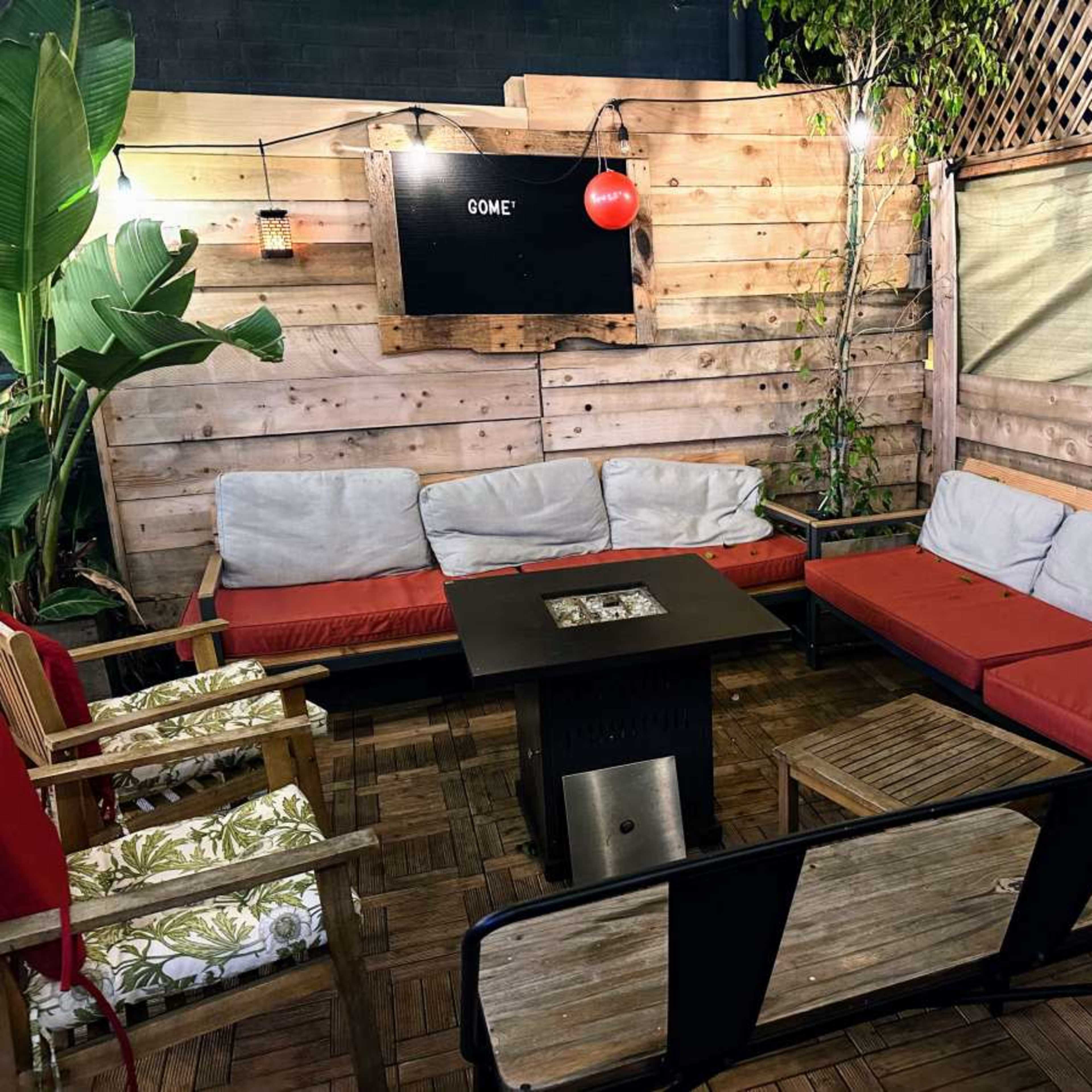 A cozy outdoor lounge area with wooden benches, cushions, and a central fire pit, surrounded by greenery and wooden walls.