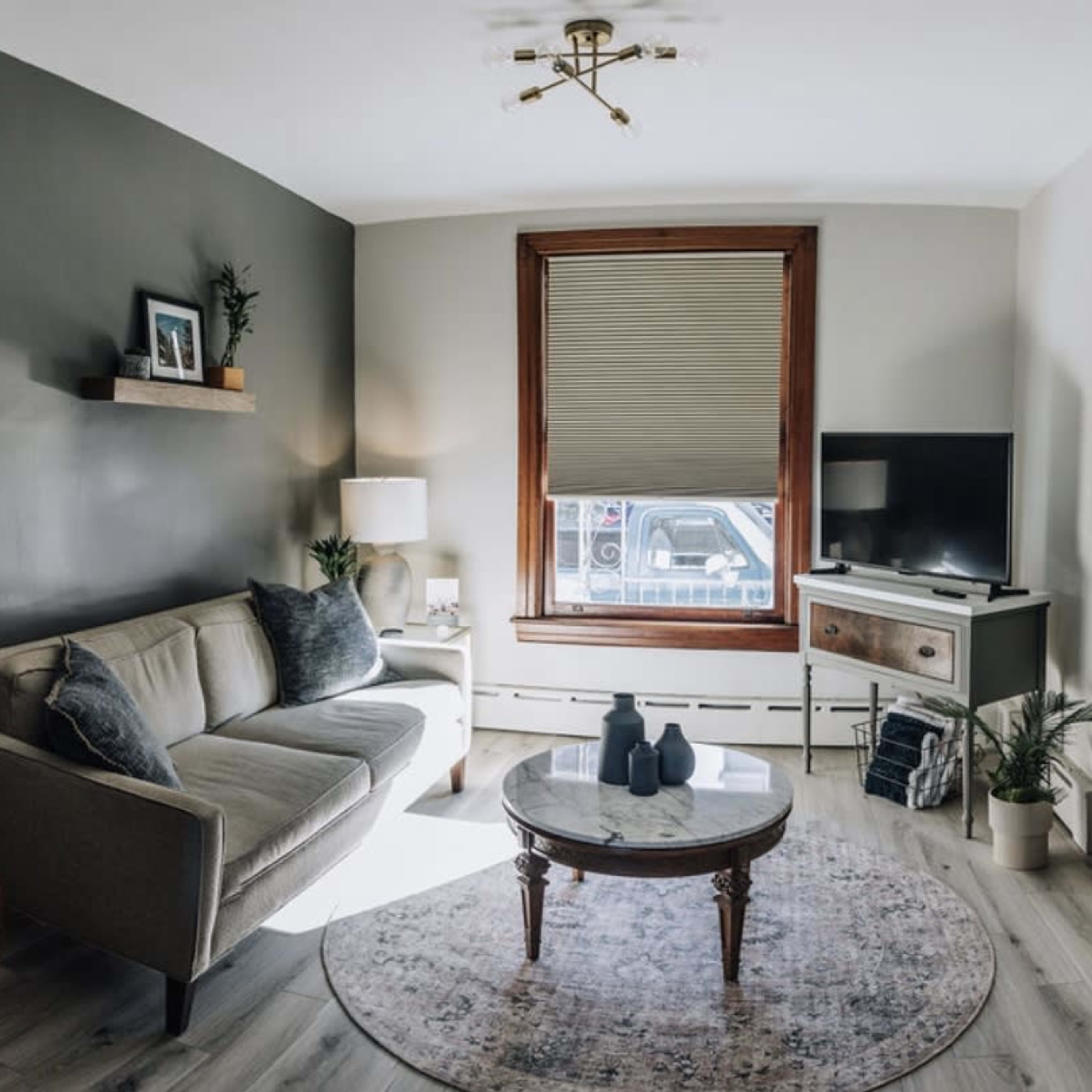 A cozy living room with a gray accent wall, a modern sofa, a round coffee table, and a television on a wooden stand, illuminated by natural light from a window.