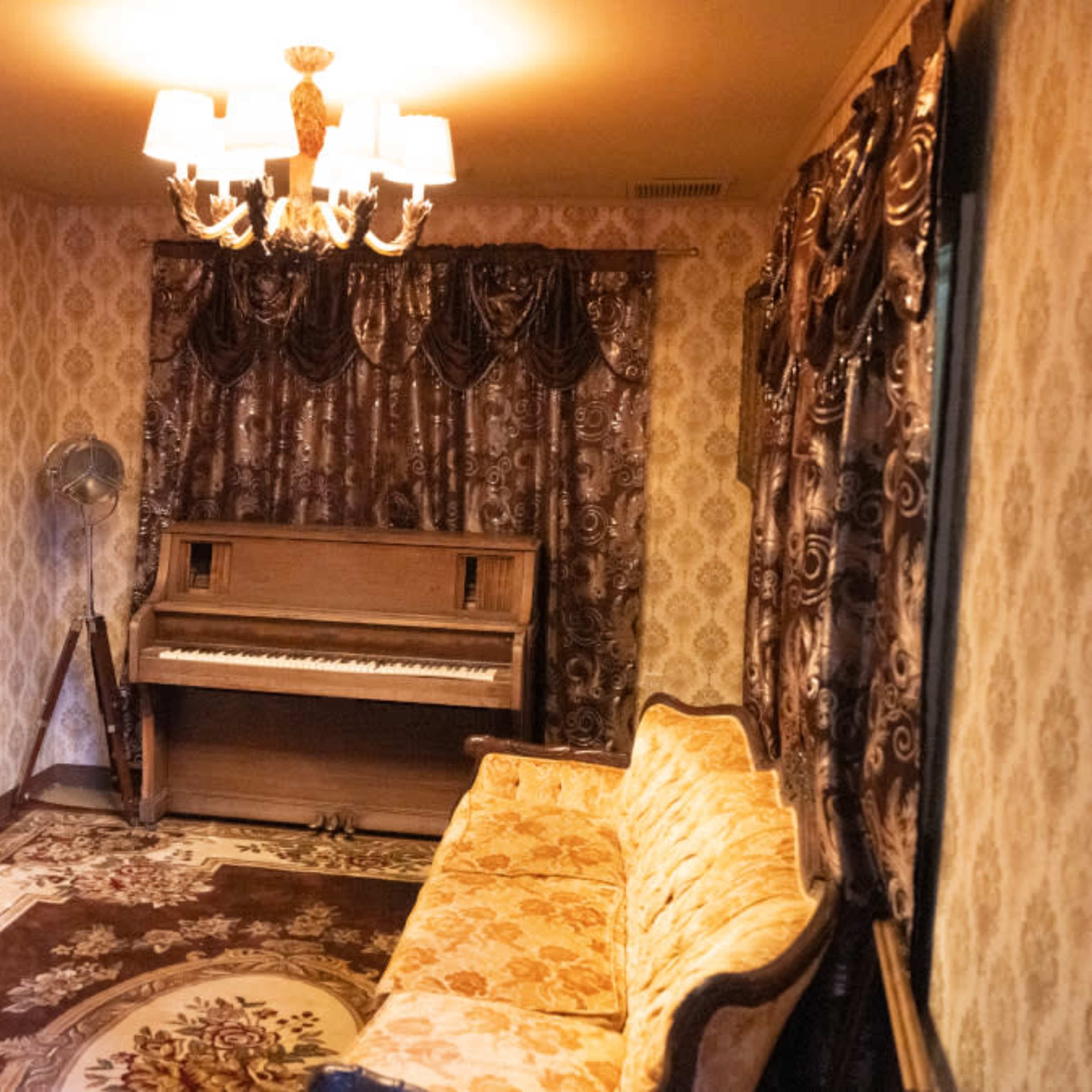 The room features a vintage piano, an ornate mirror, and a decorative sofa, all set against patterned wallpaper and drapes.
