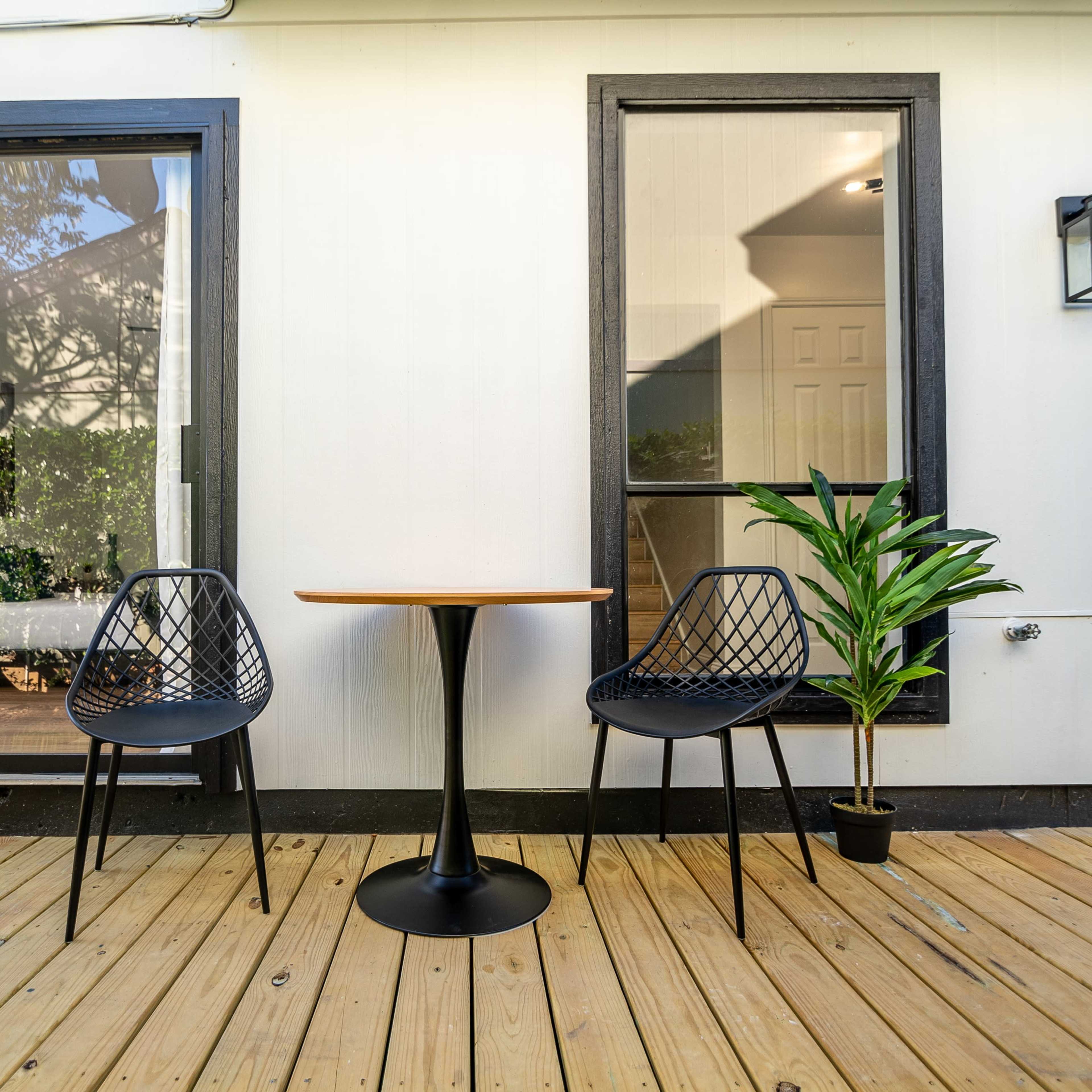 A small outdoor seating area with two black chairs and a round table on a wooden deck, adjacent to a house with large windows.
