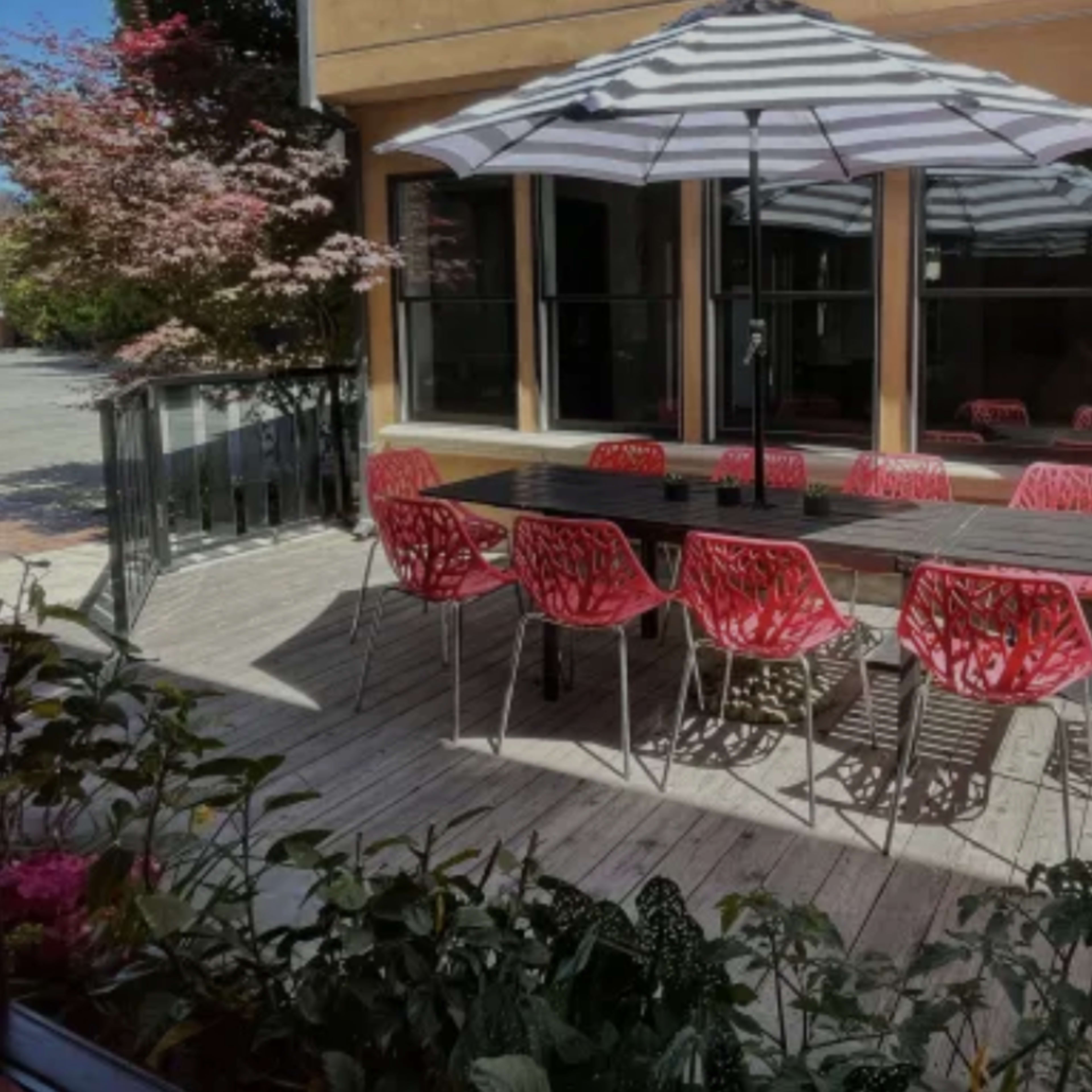 A patio features a large wooden table surrounded by red chairs, with a striped umbrella providing shade.