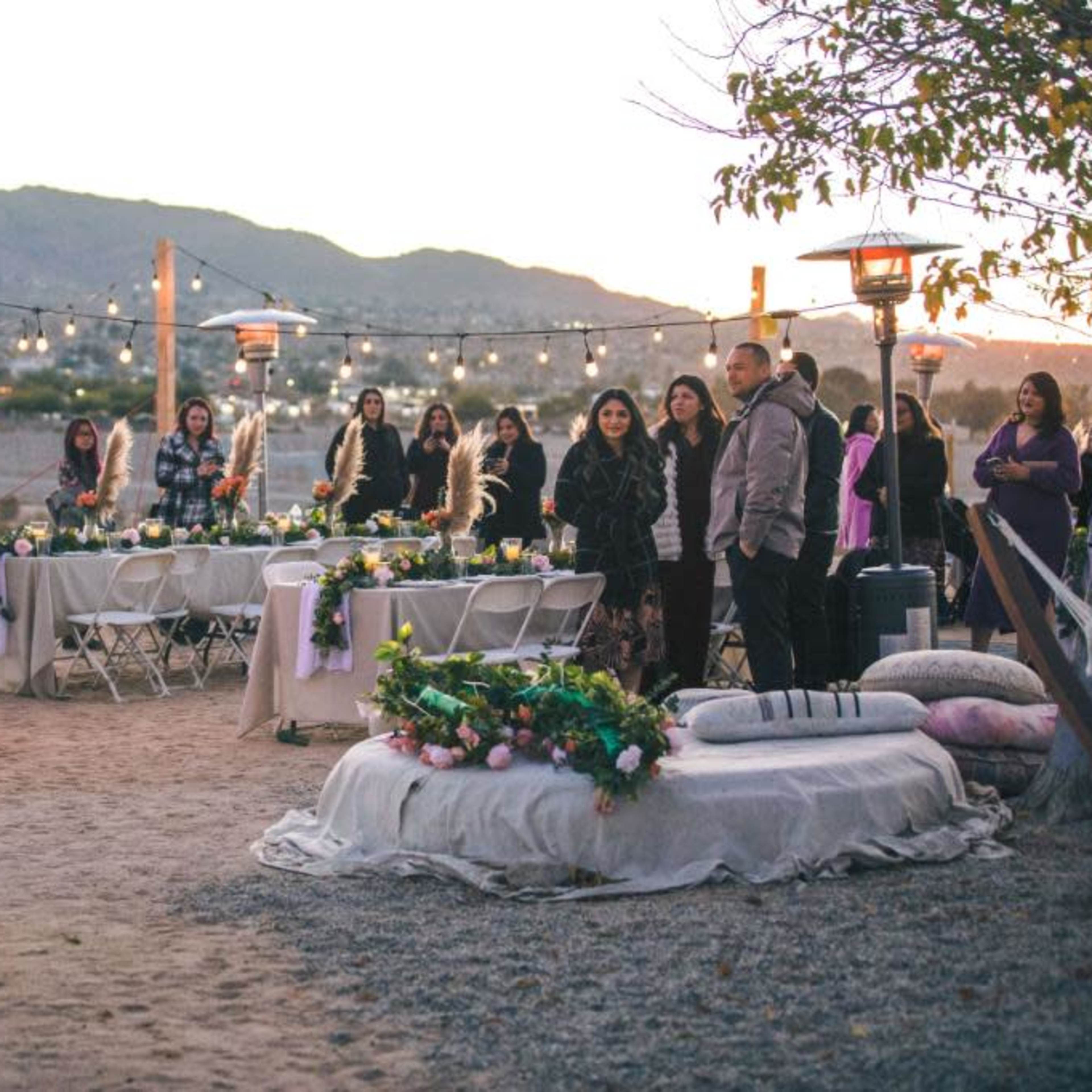 A bride stands in front of guests at an outdoor wedding reception decorated with lights and floral arrangements at sunset.