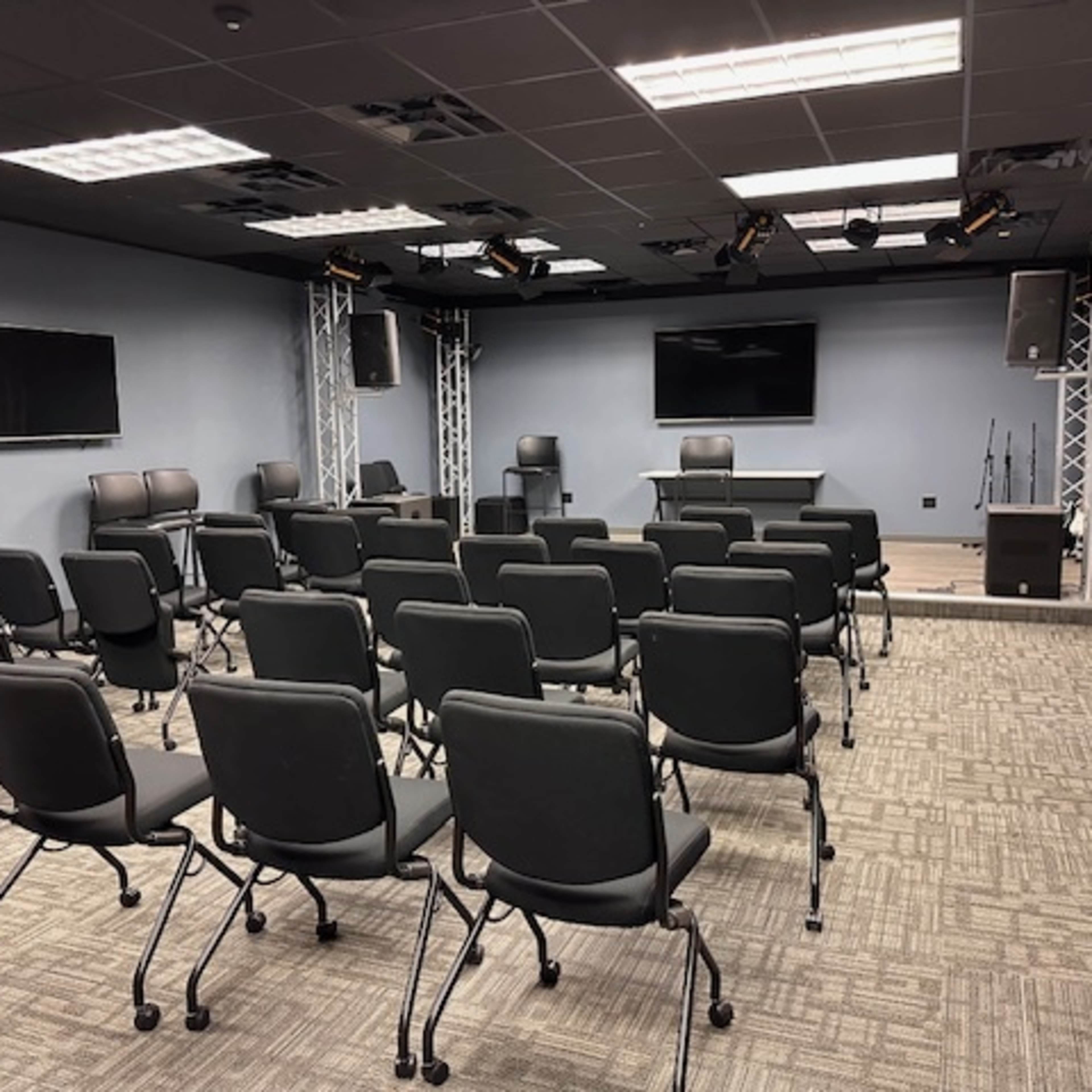A modern conference room set up with rows of black chairs, a presentation area at the front, and audio-visual equipment.