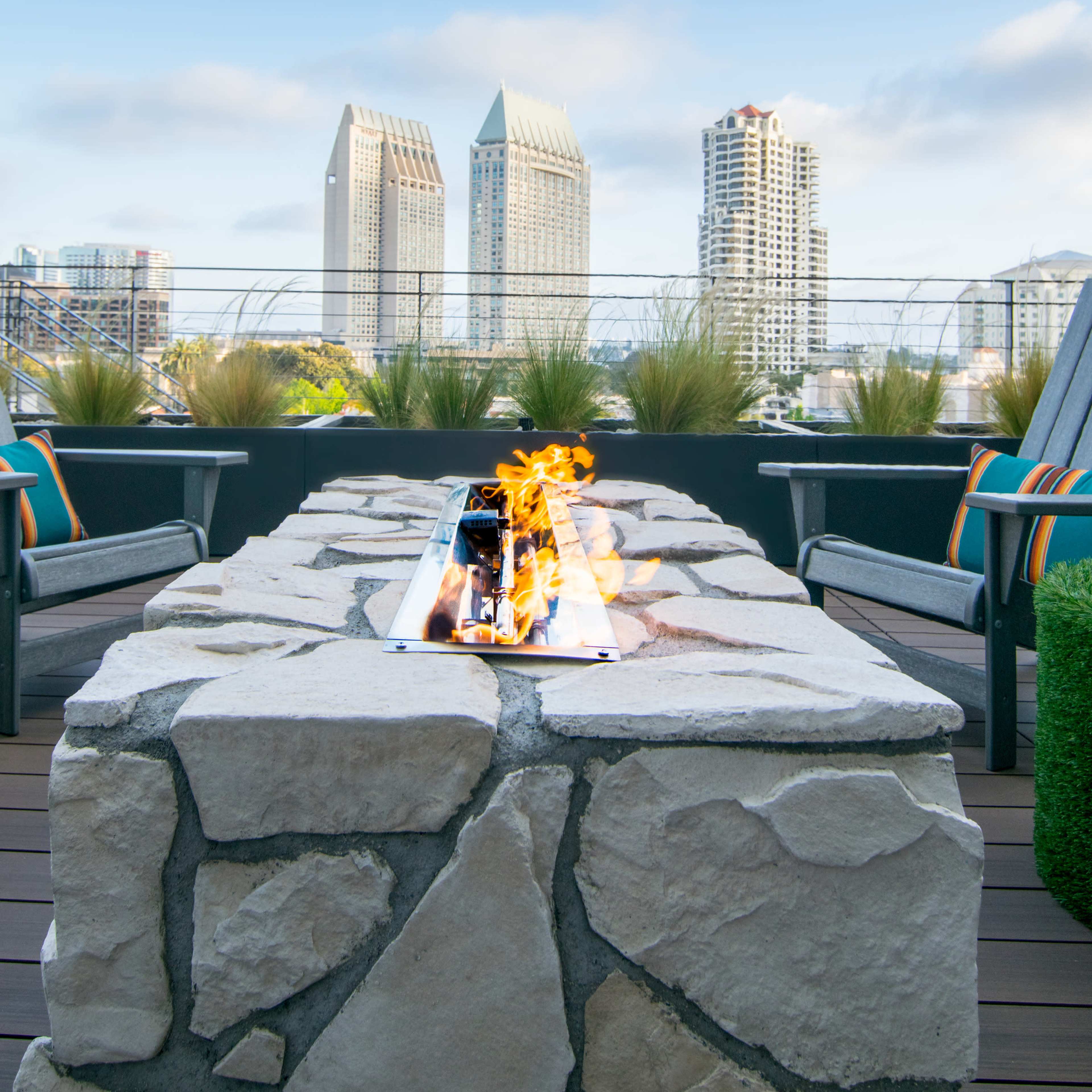A rooftop patio features a stone fire pit with flames, surrounded by chairs and offering a view of tall buildings against a clear blue sky.
