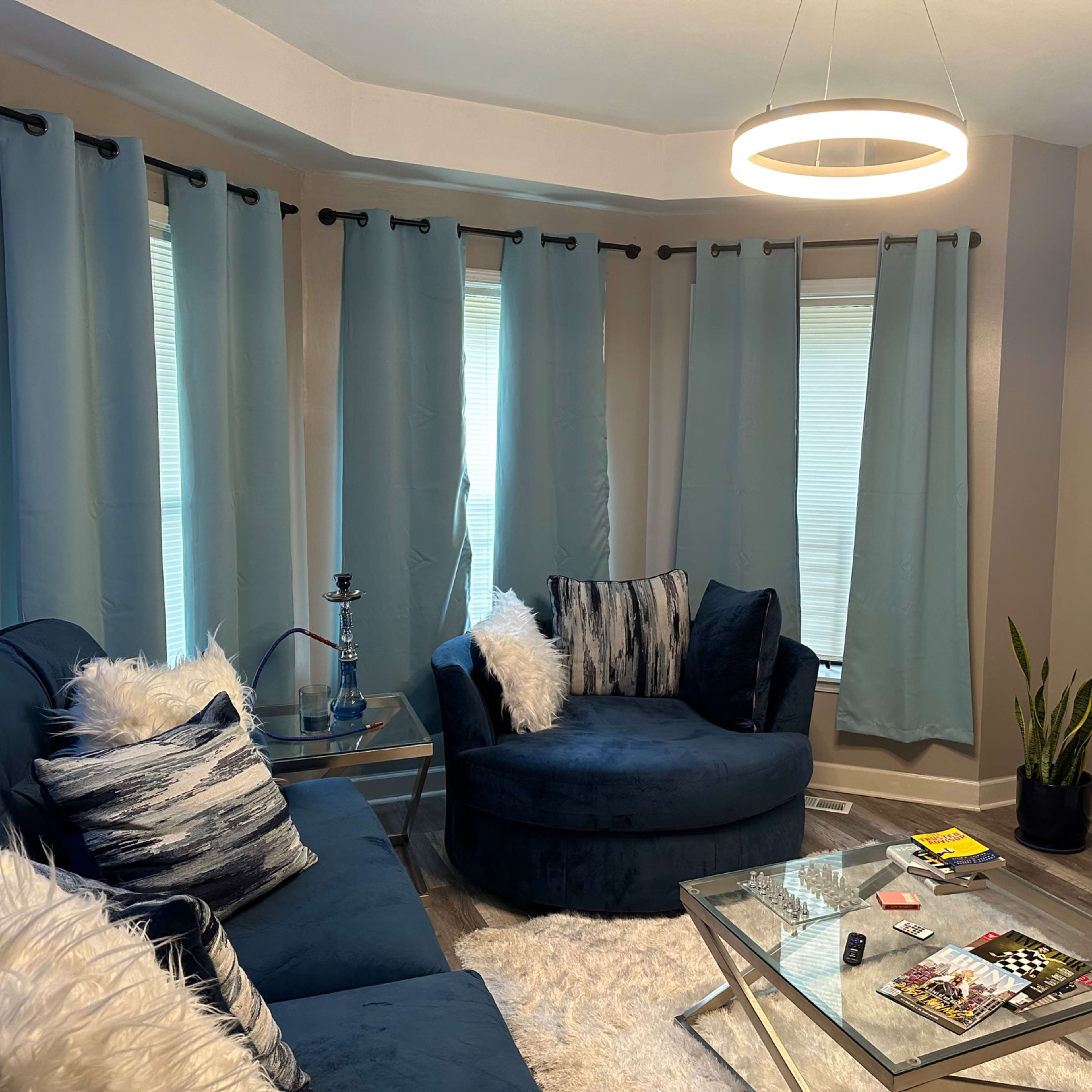 A living room with blue curtains, a round chair, a sectional sofa, a glass coffee table, and a white rug.