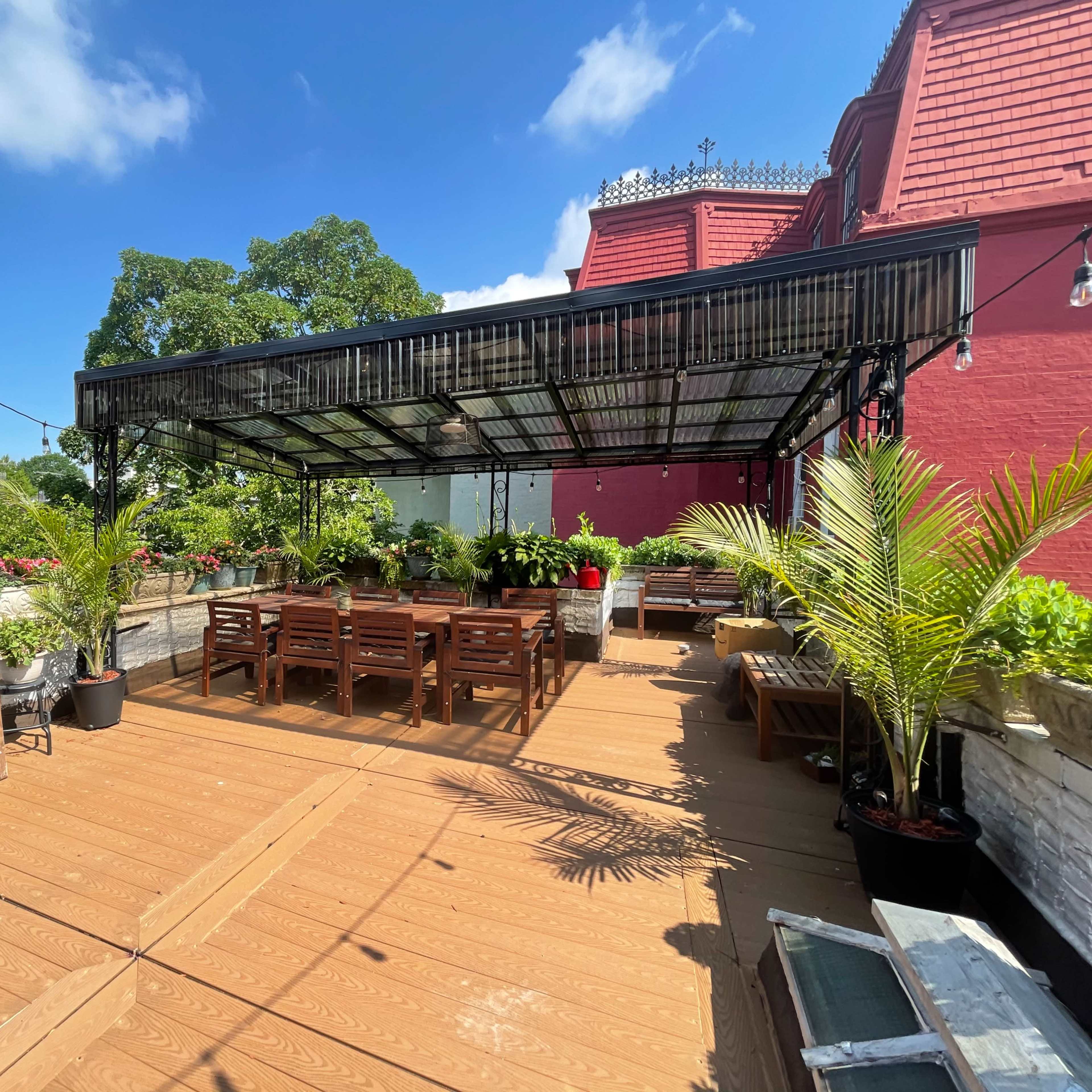 A rooftop terrace with wooden seating, greenery, and a large shaded area under a pergola.