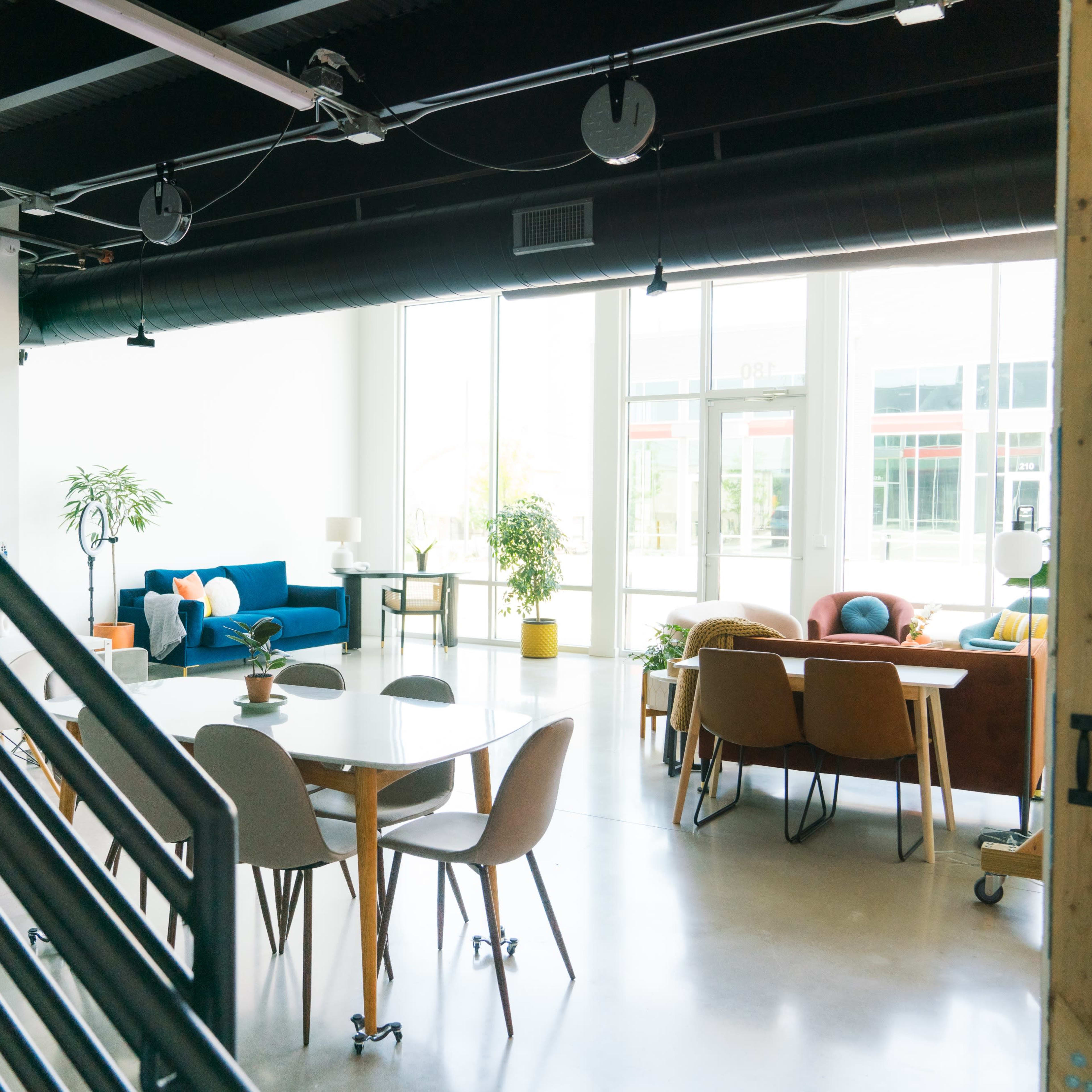 A spacious, modern interior with a dining table surrounded by chairs, a staircase, and various seating areas, illuminated by large windows.