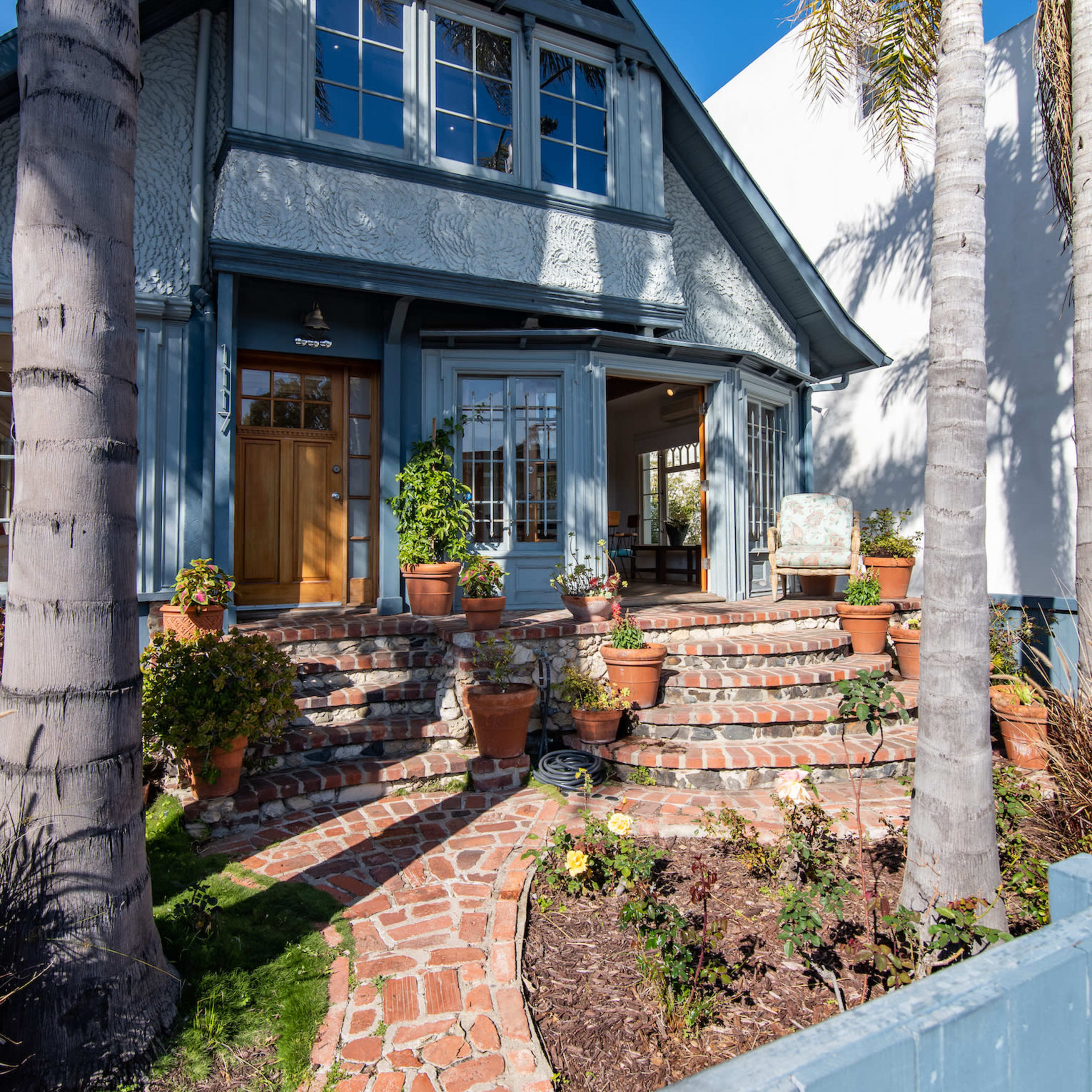 A blue bungalow with a landscaped front yard is framed by palm trees and features a brick pathway leading to the entrance.