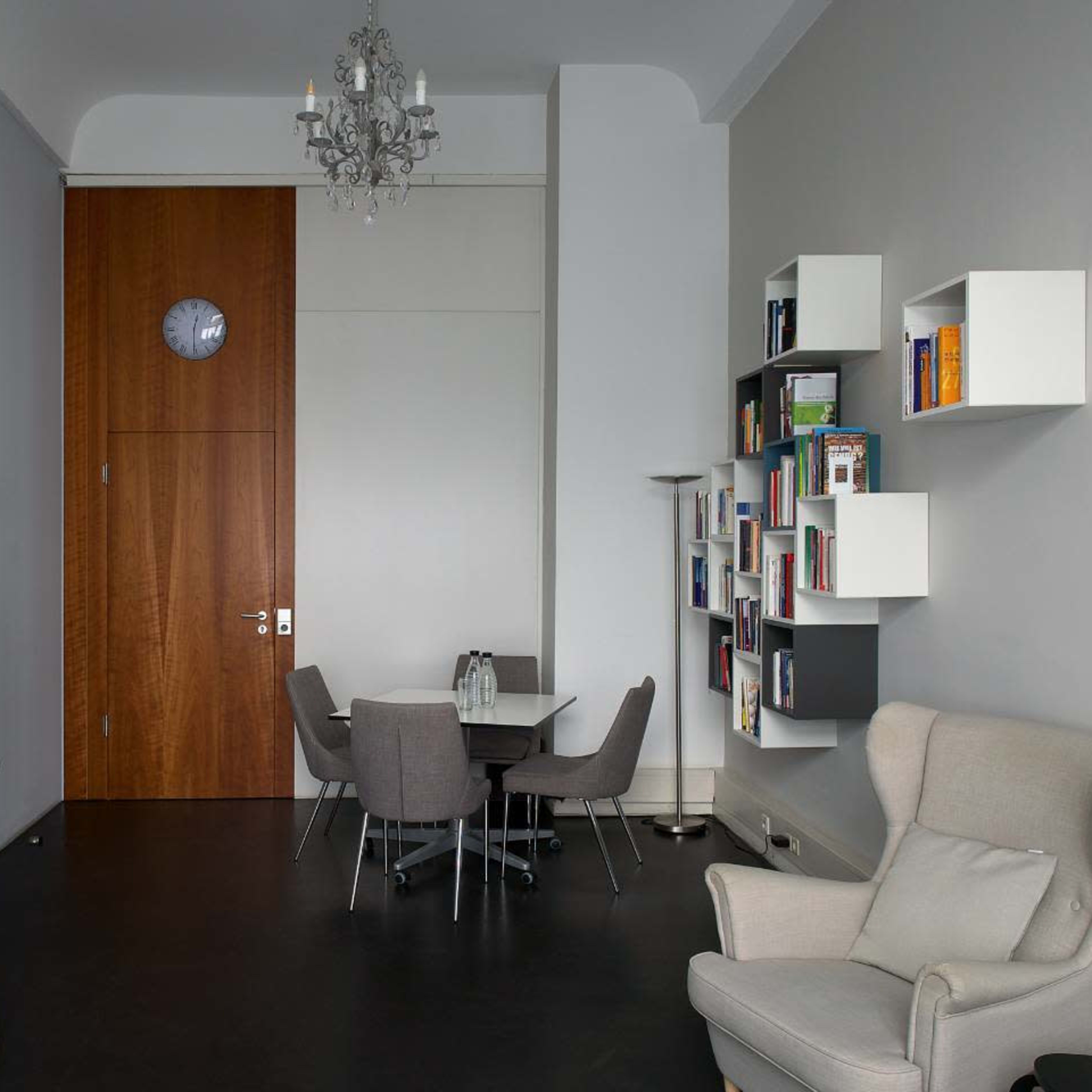 A modern office space featuring a round table with four chairs, a bookshelf, a whiteboard on an easel, and a wood-paneled door with a clock above it.