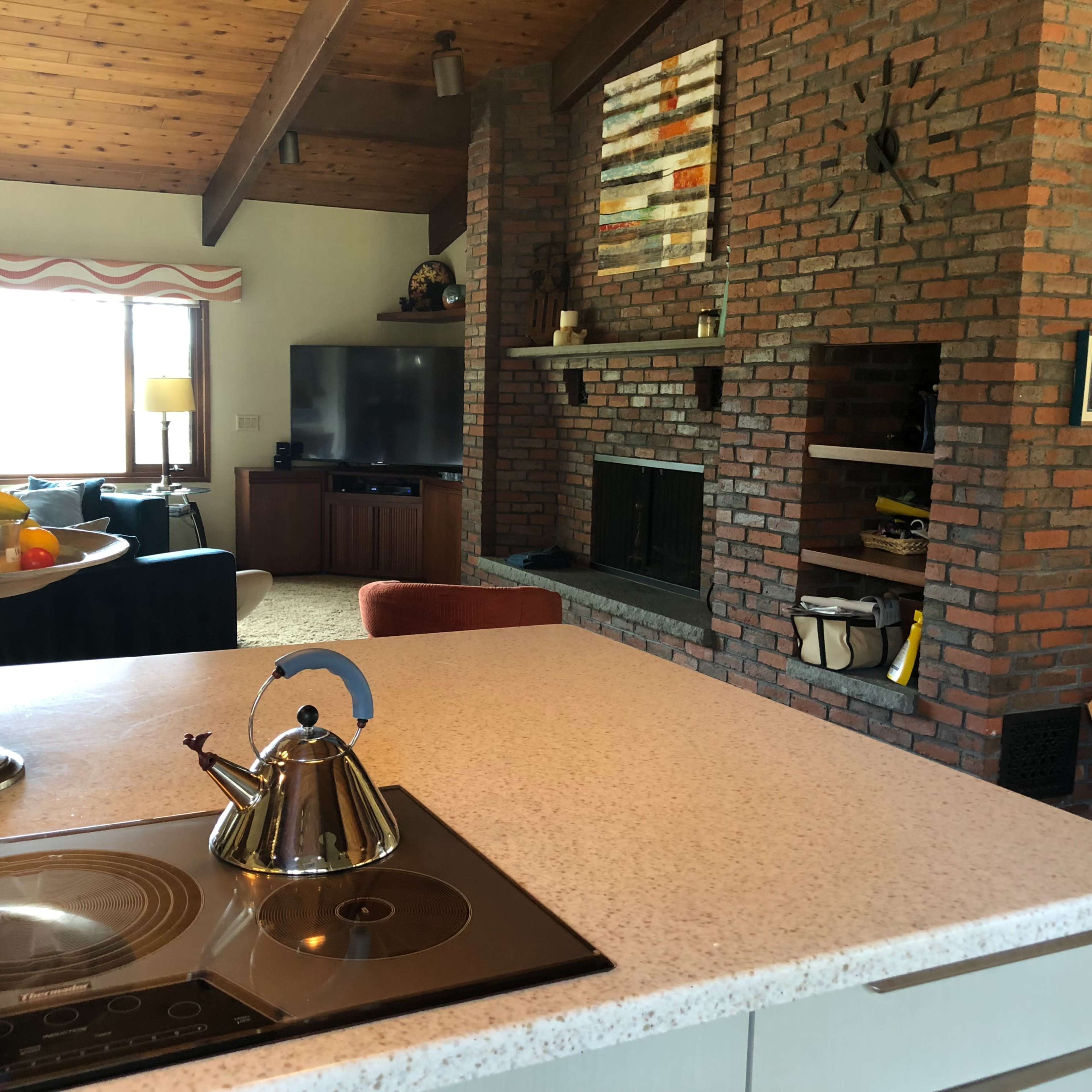A modern kitchen with a countertop, featuring a kettle and a bowl of fruit, adjacent to a living room with brick walls and a television.
