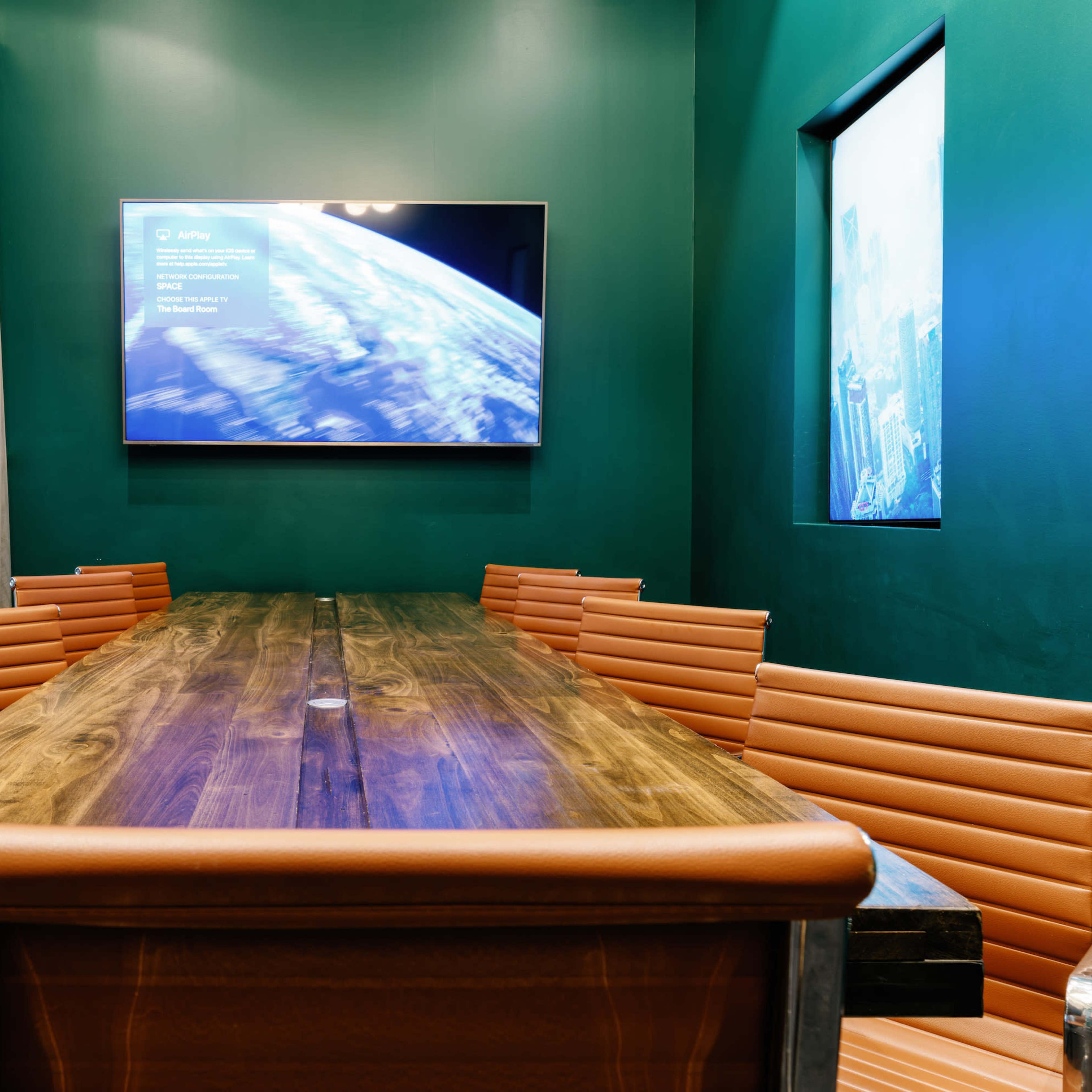 A conference room featuring a long wooden table surrounded by orange striped chairs, with a television mounted on a green wall displaying information.