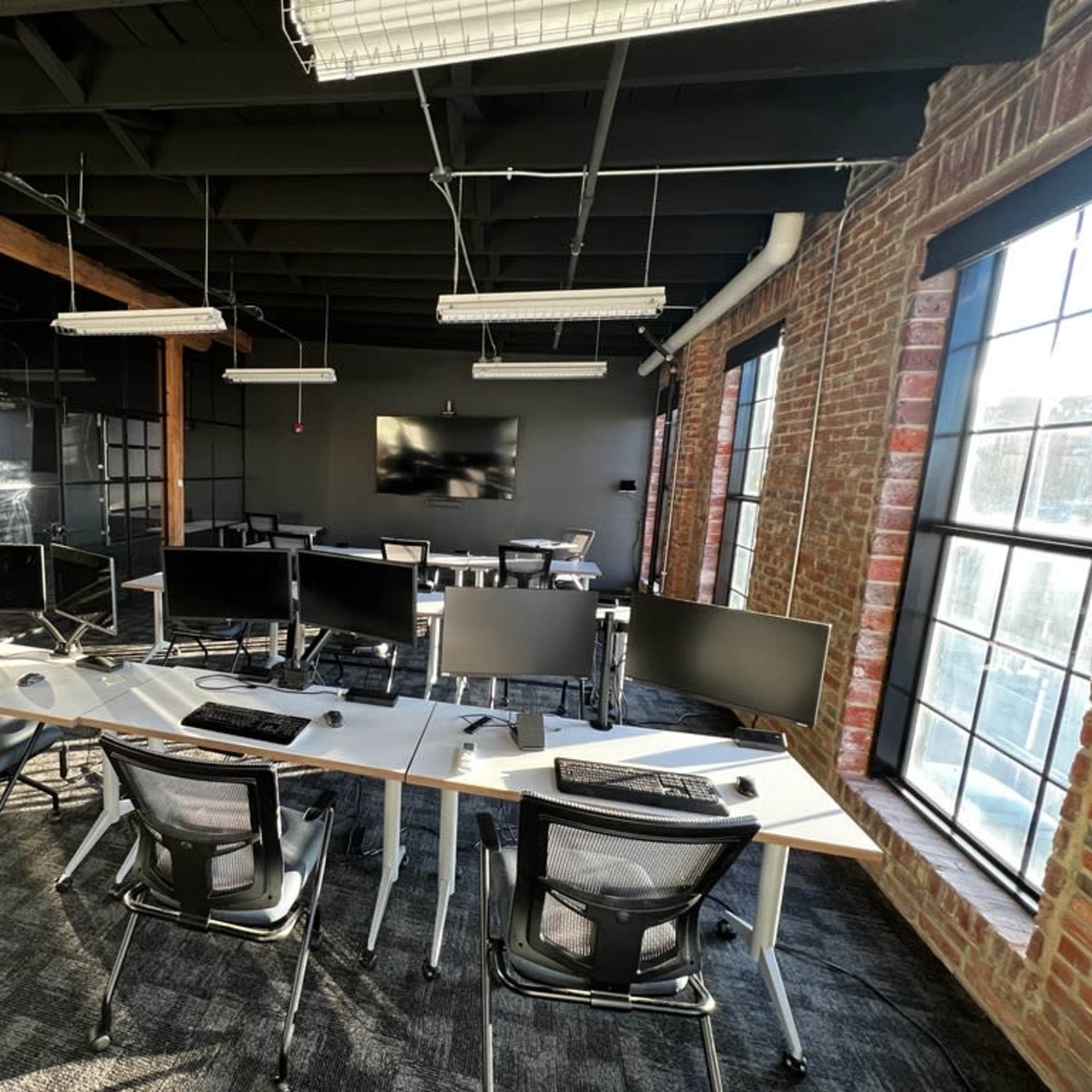 A modern office space featuring multiple workstations with computer monitors and a conference area with a large screen, surrounded by large windows and exposed brick walls.