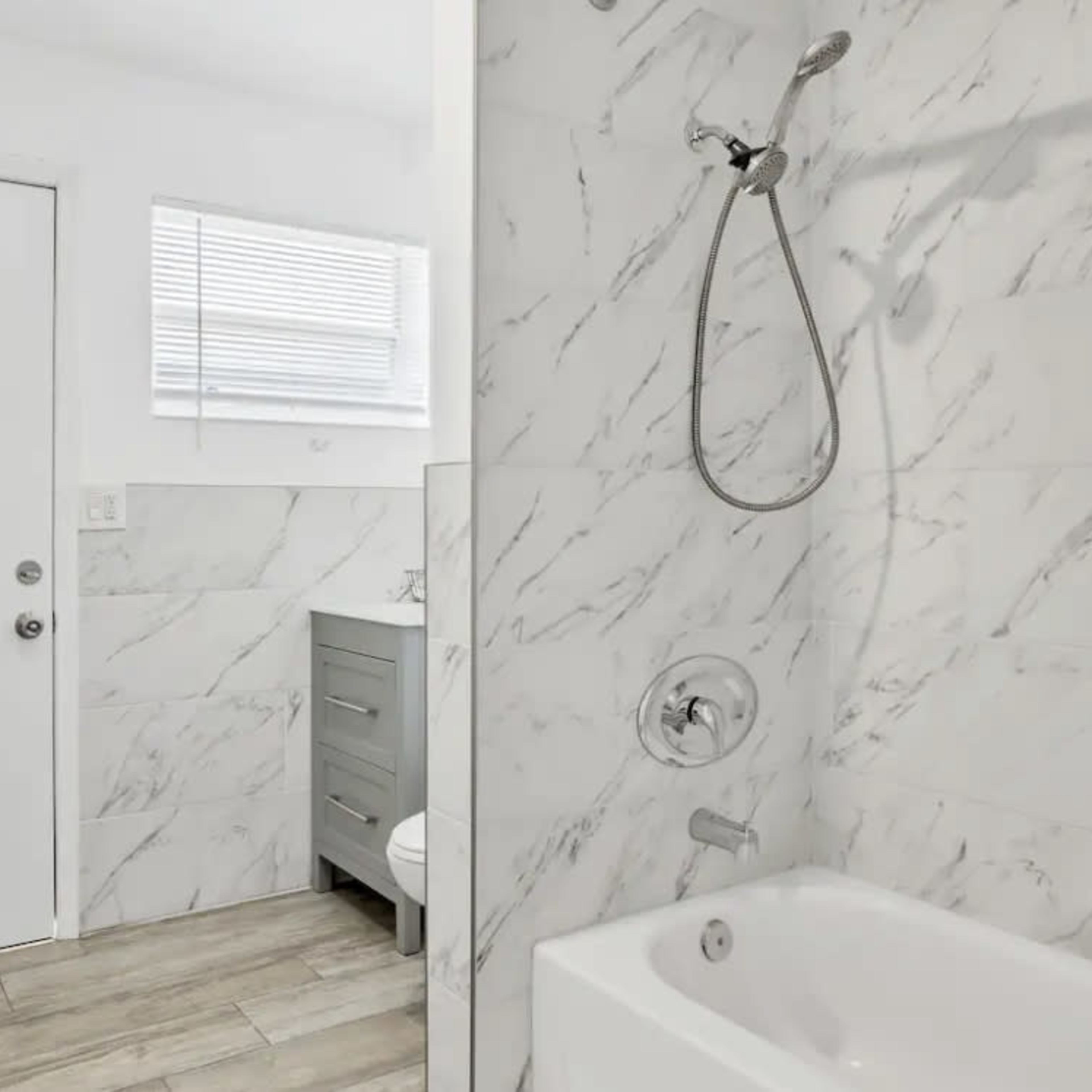 A bathroom featuring a marble-tiled shower area, a bathtub, and a gray vanity near a door and window.