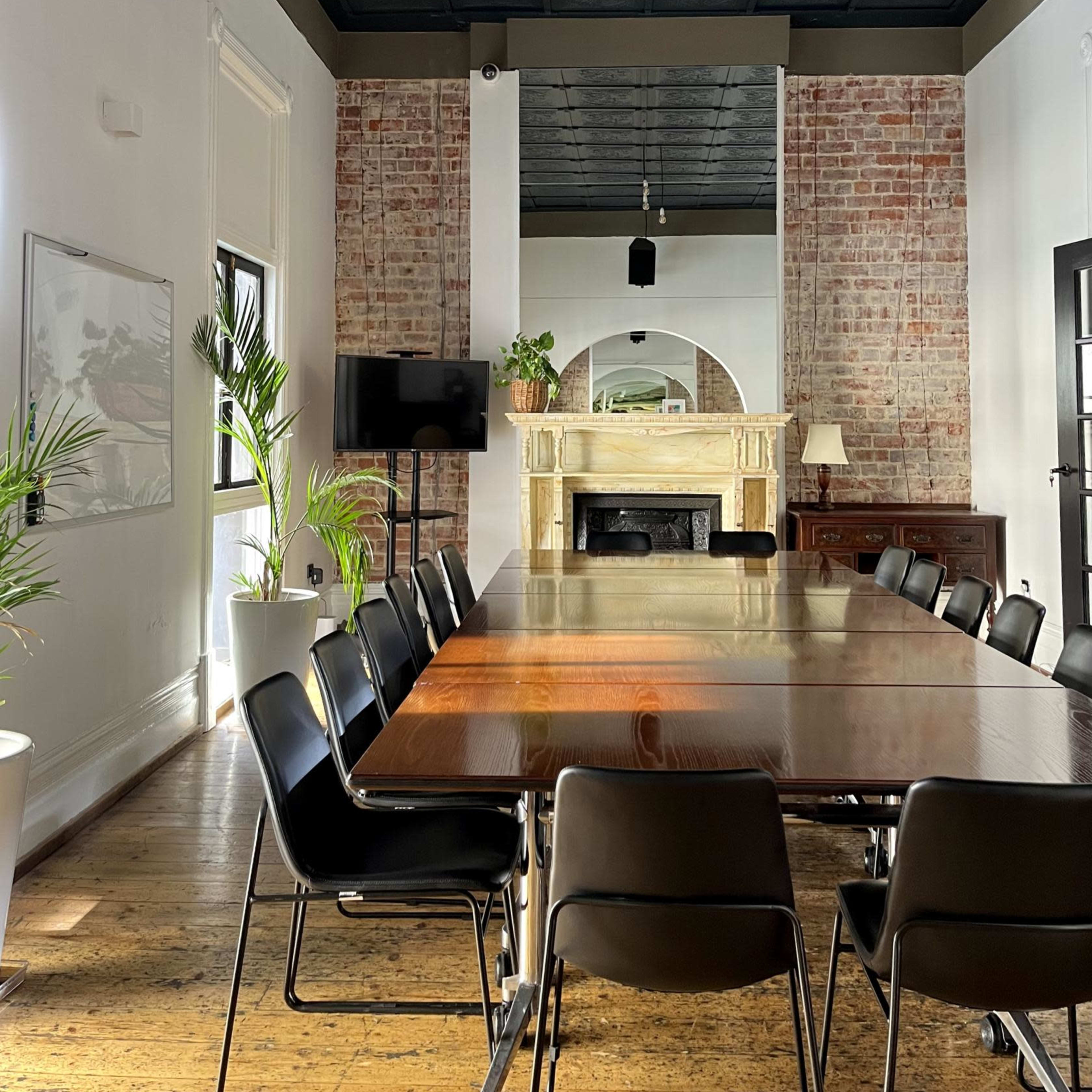 A long wooden table surrounded by black chairs is set in a spacious room with exposed brick walls, a fireplace, and large windows allowing natural light to enter.