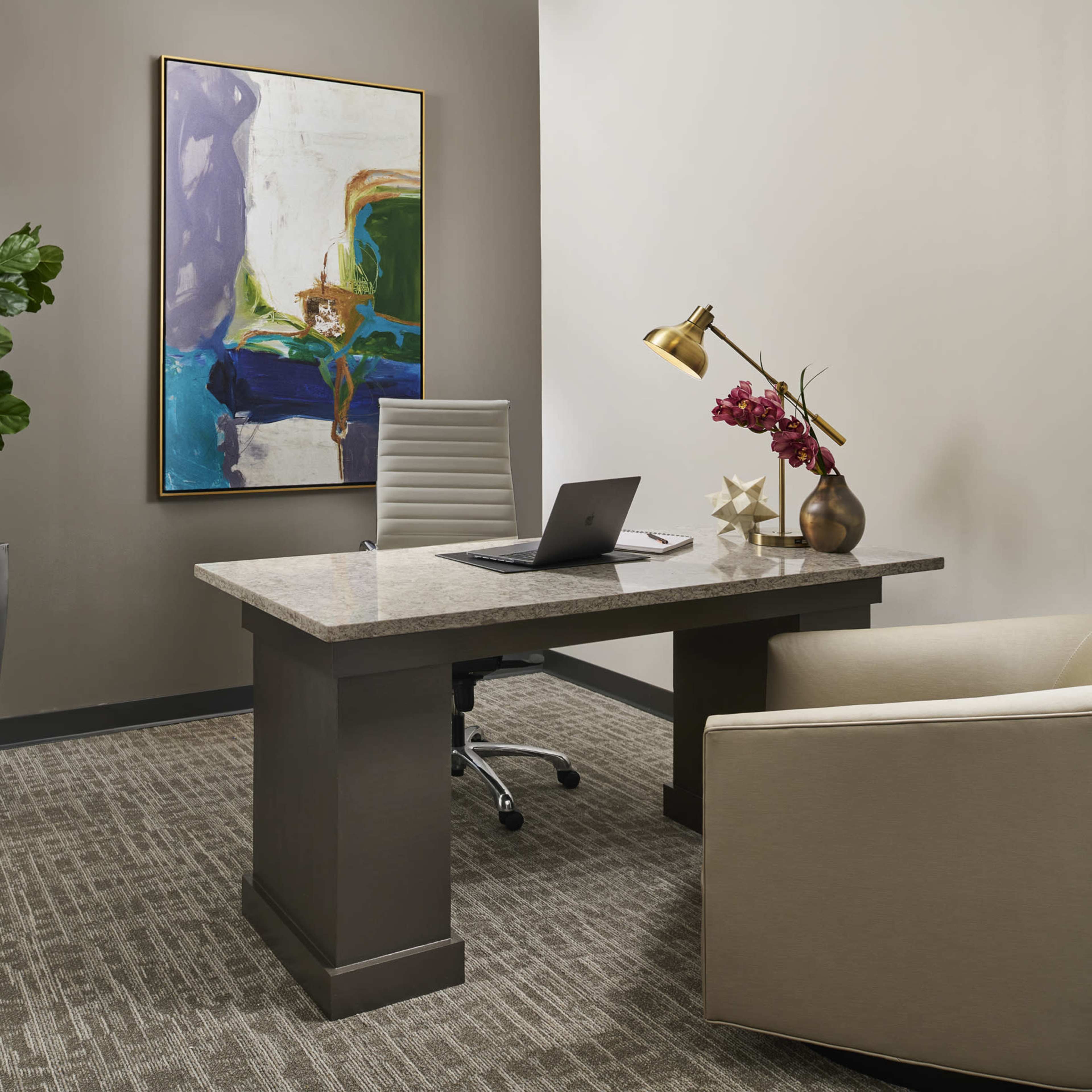 An office features a sleek desk with a laptop, a comfortable chair, a potted plant, and a large piece of artwork on the wall.