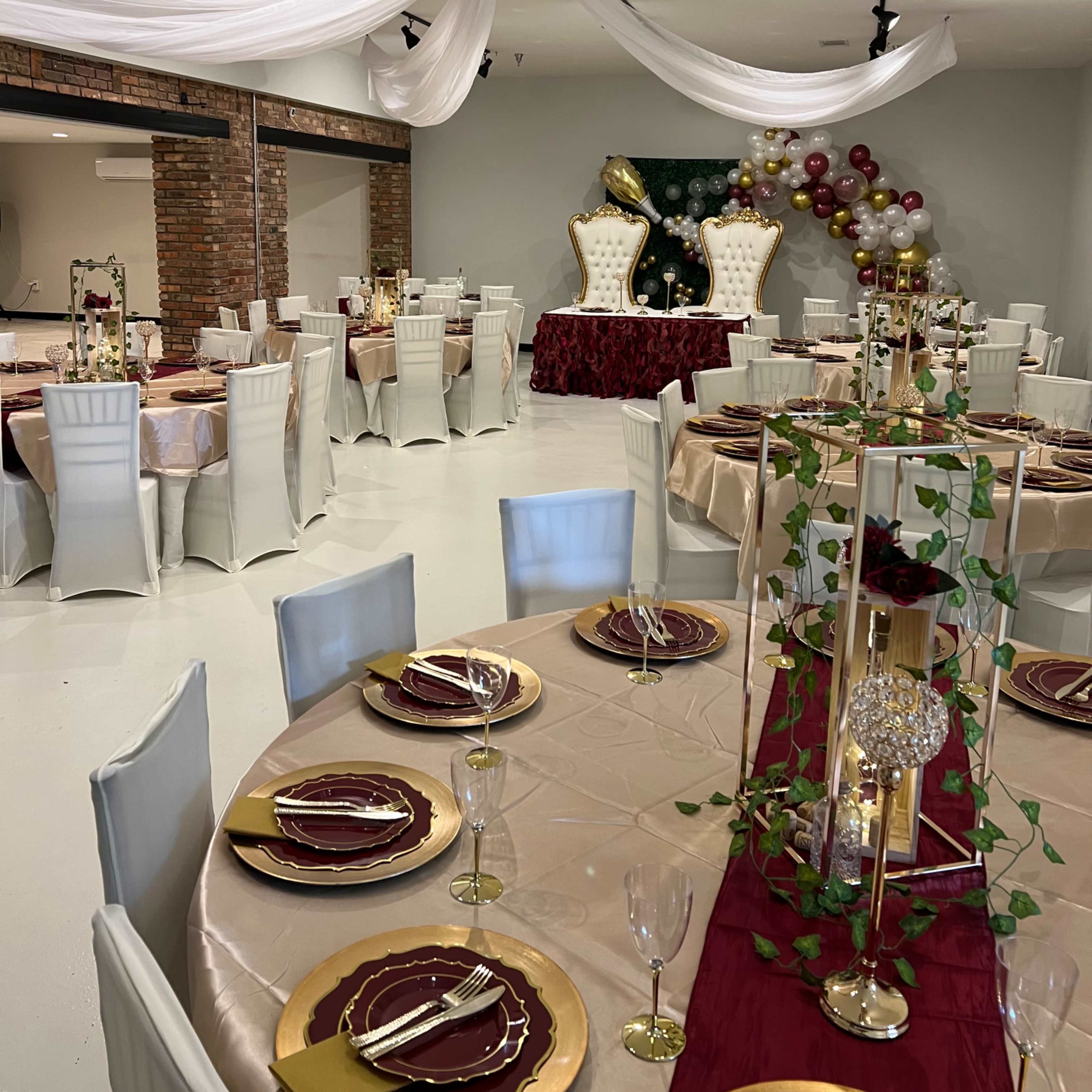 A elegantly decorated event space with round tables set for dining, white chairs, and a backdrop featuring a large floral arrangement with balloons.