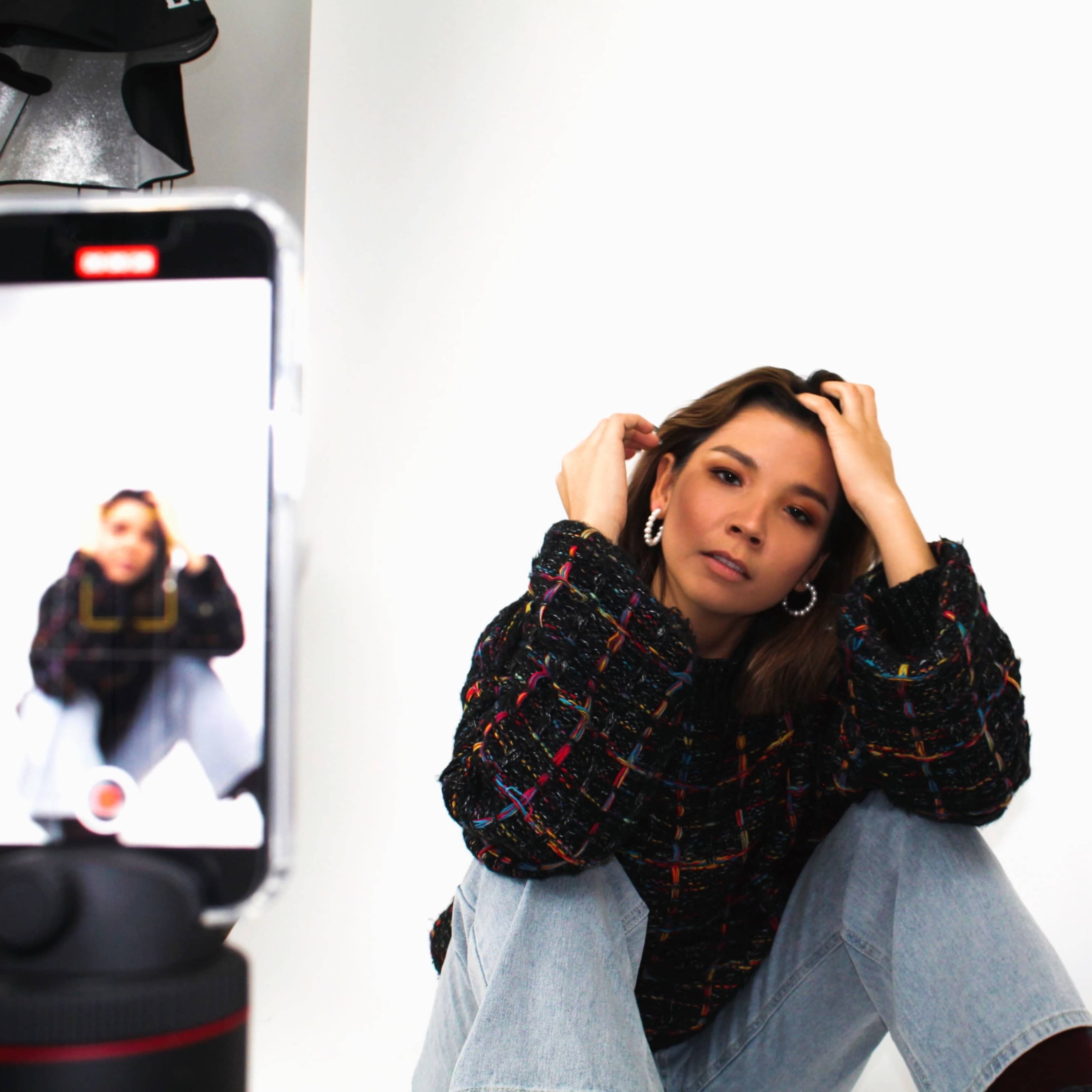 A woman in a colorful sweater sits on the floor while posing for a photo taken with a smartphone.