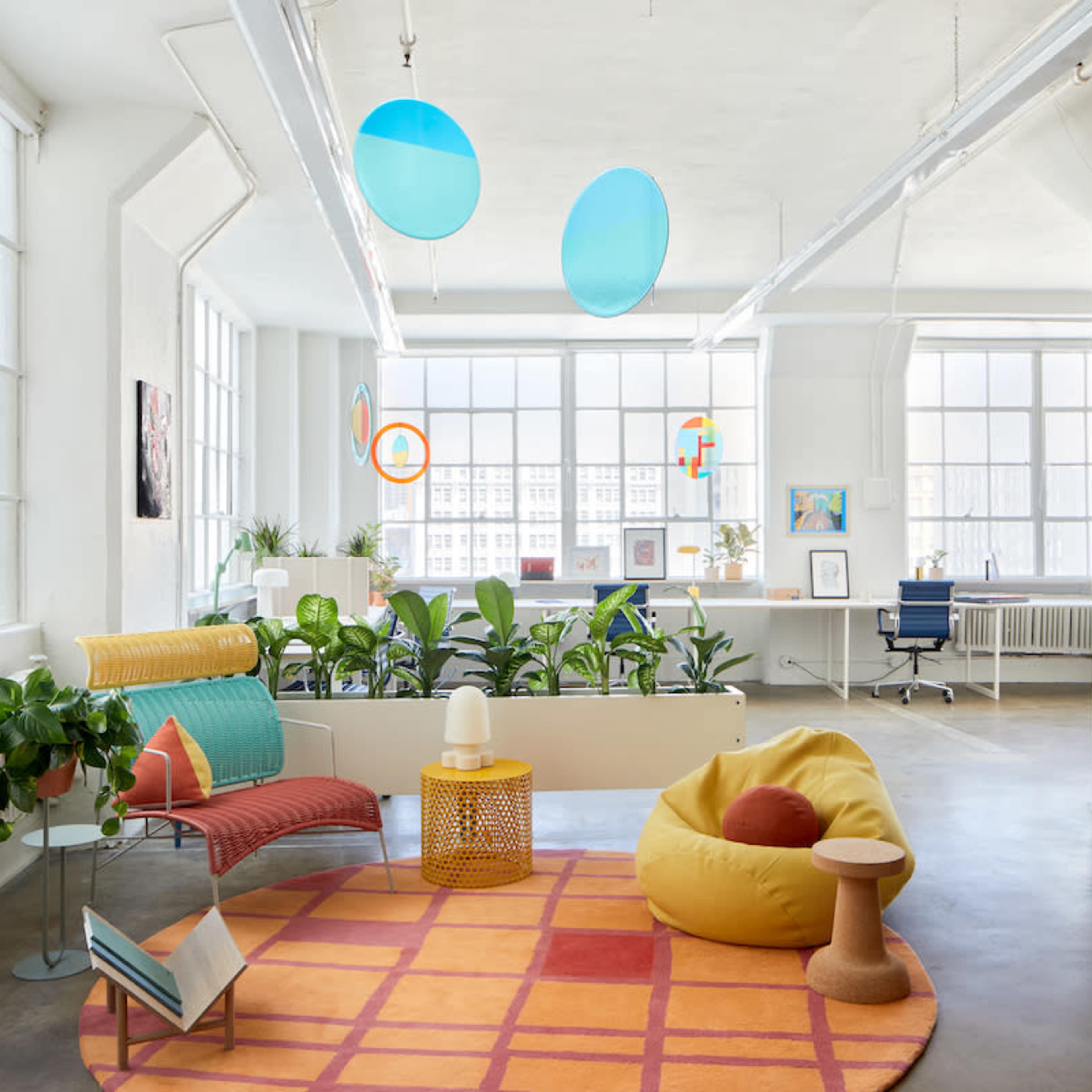 A brightly lit modern office space featuring large windows, a variety of plants, colorful furniture, and a bookshelf against a textured wall.