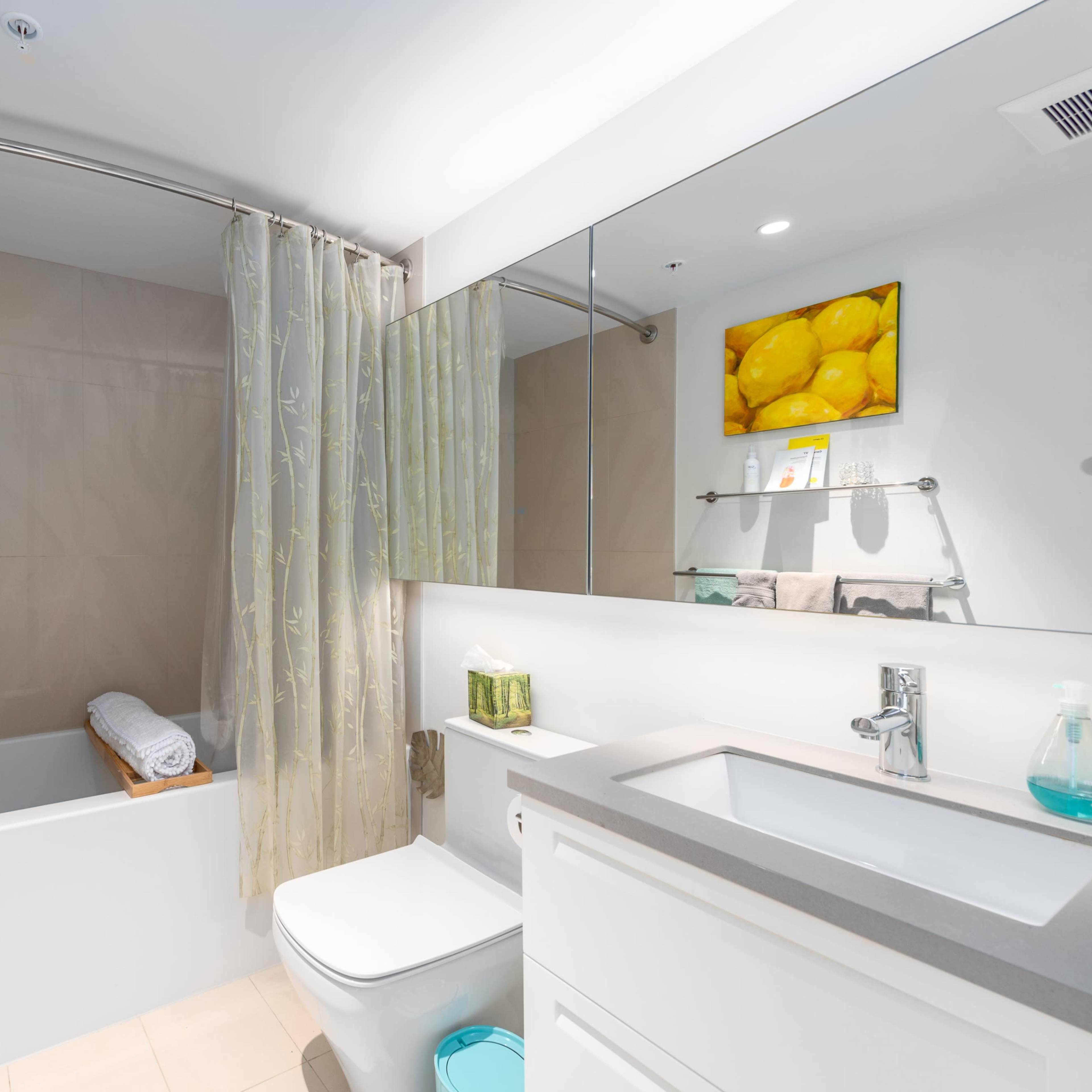 A clean bathroom features a bathtub with a shower curtain, a modern sink with a gray countertop, and lemon-themed artwork on the walls.