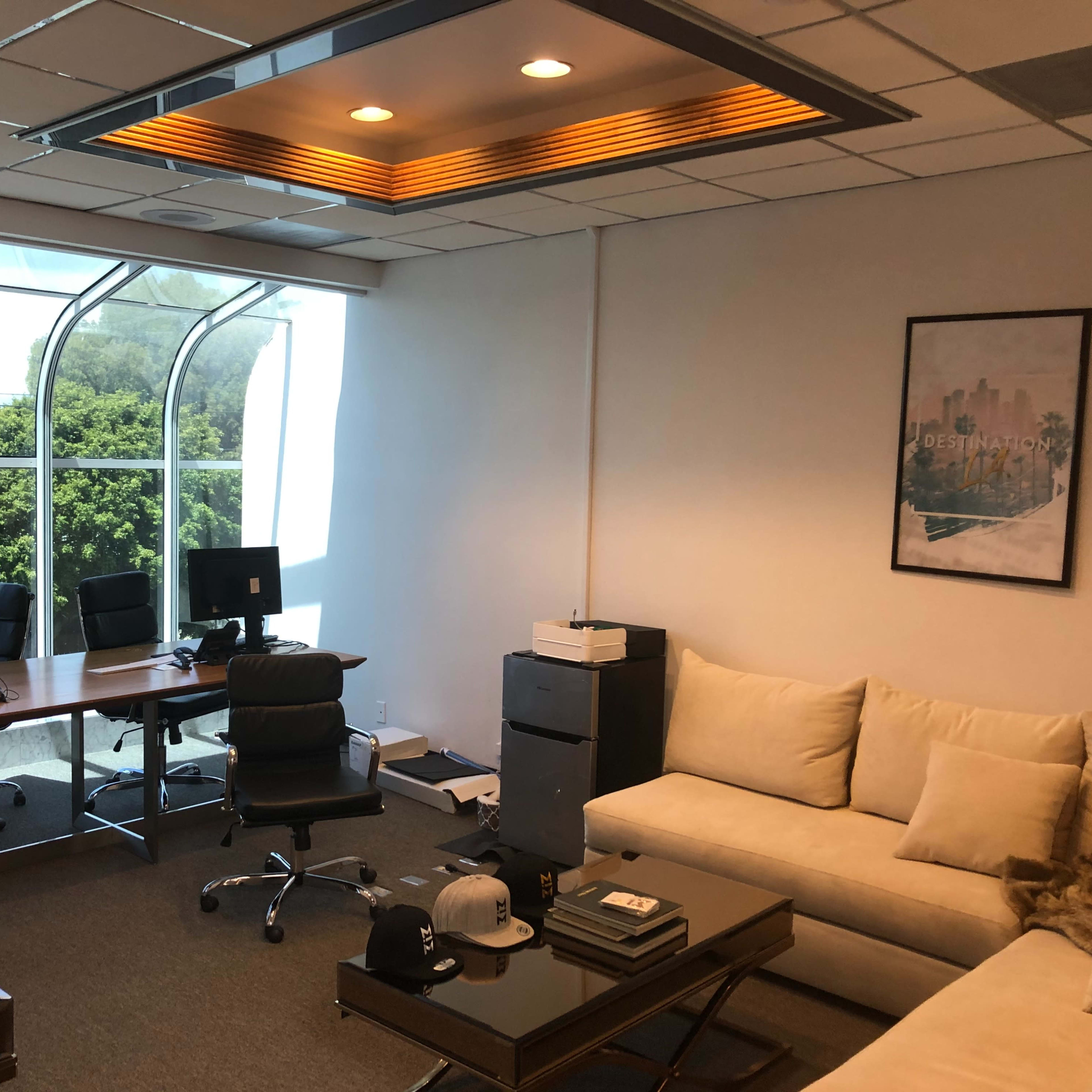 An office space featuring a large window, a sectional sofa, a coffee table, and a workspace with desks and office equipment.