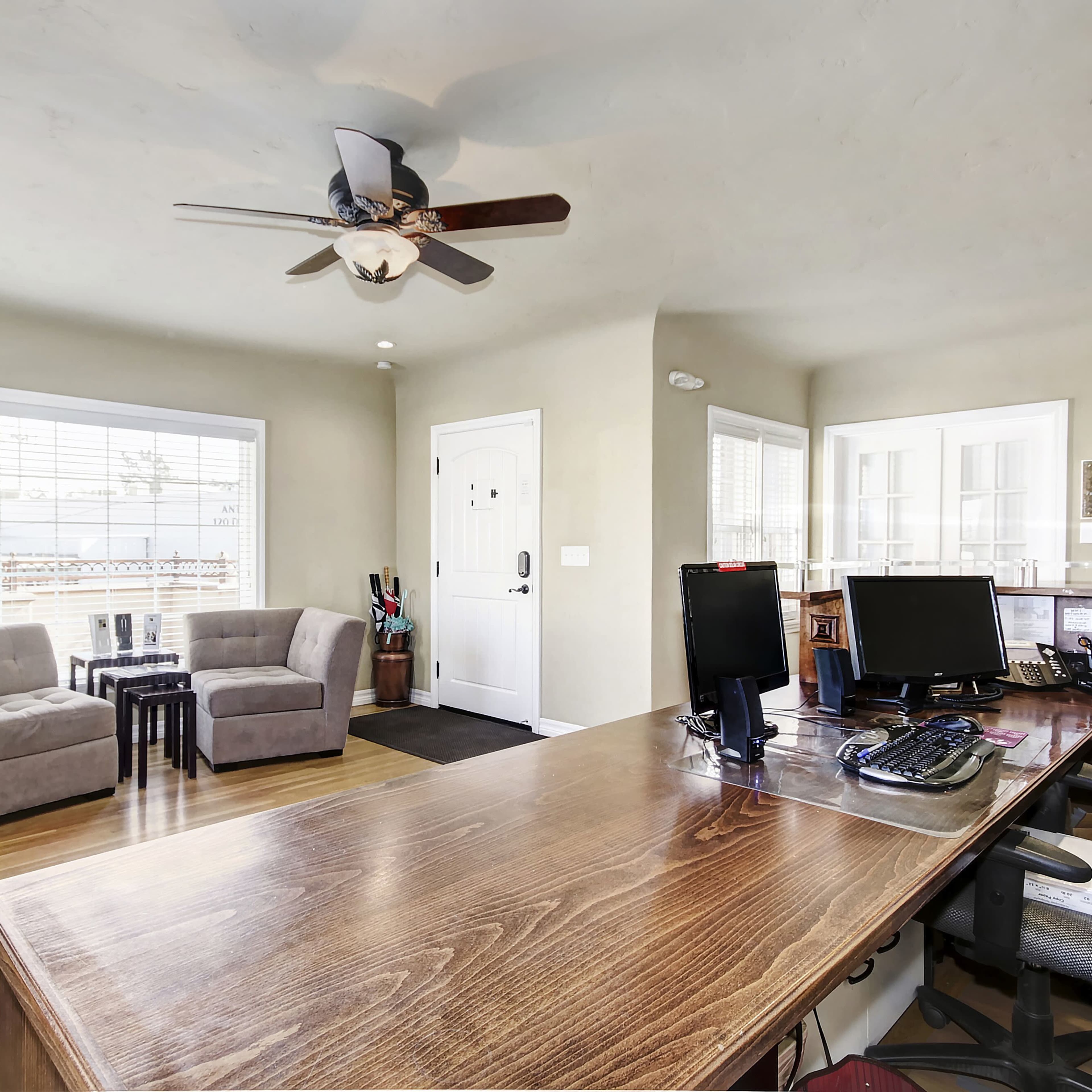A home office with a wooden desk, two chairs, and a patio door, featuring a ceiling fan and a potted plant in the corner.