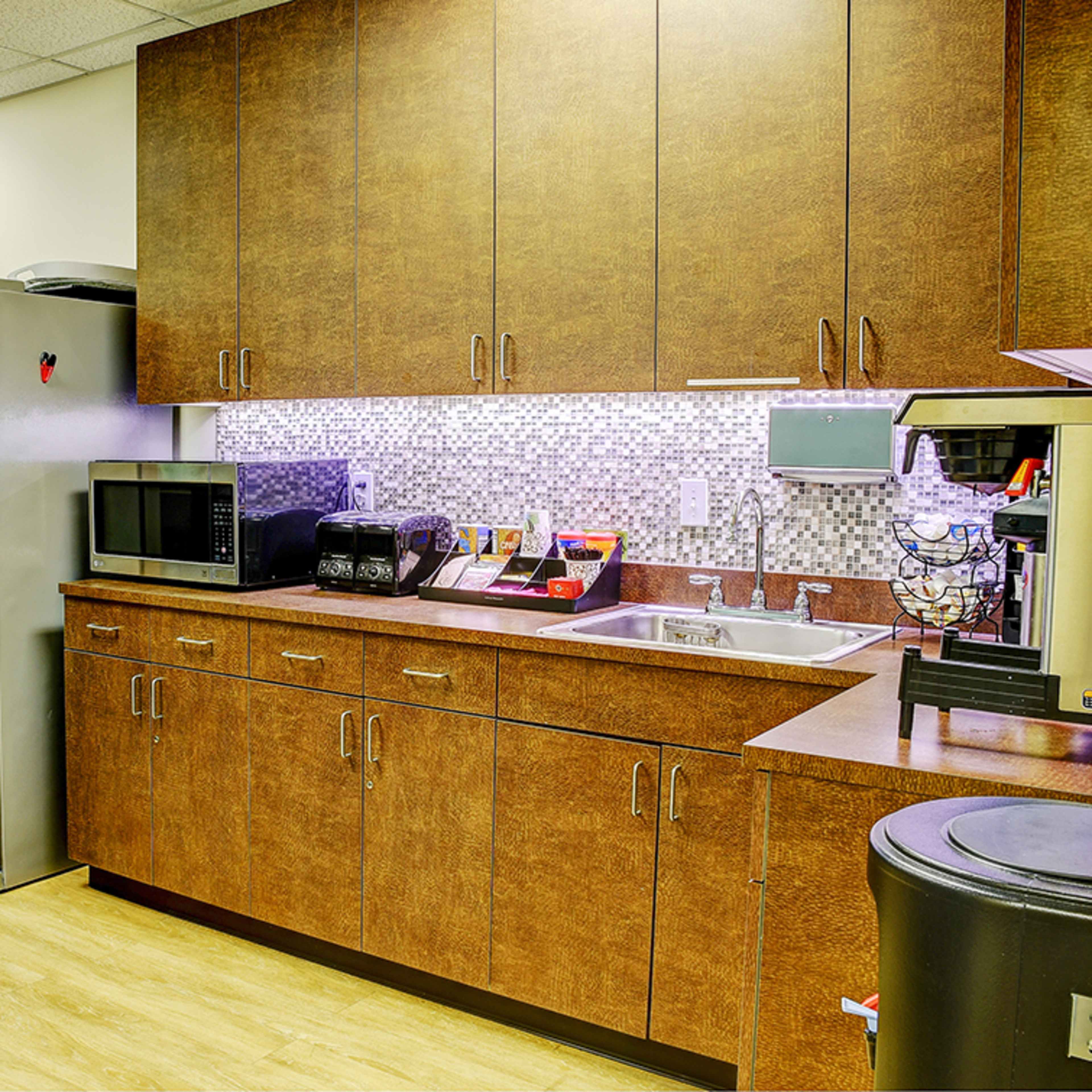 A kitchen area featuring a stainless steel refrigerator, a vending machine, a microwave, a coffee maker, and wooden cabinetry with a tiled backsplash.