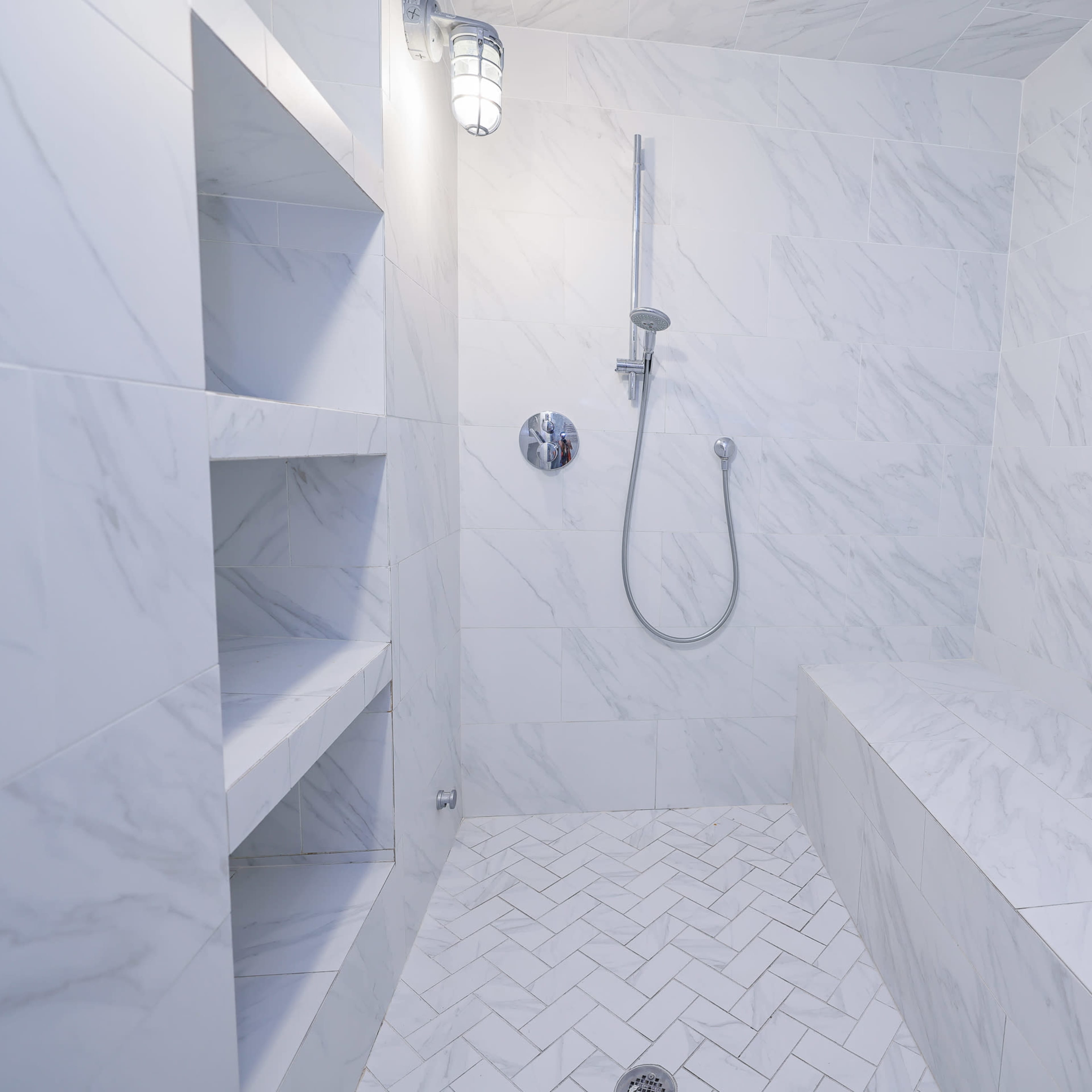 A modern shower stall with marble walls, a handheld showerhead, and built-in shelving.