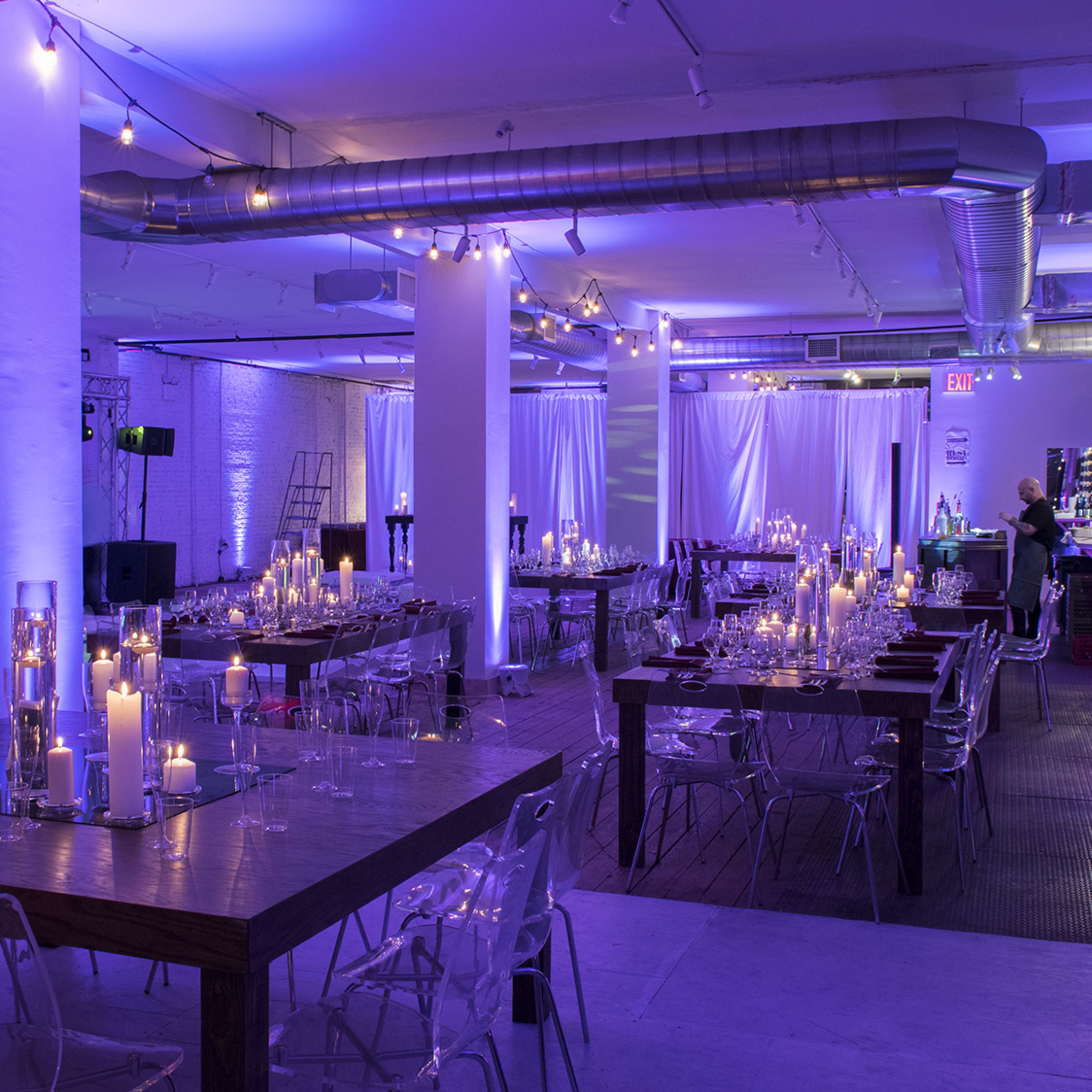 A spacious venue features several long wooden tables set for an event, illuminated by soft purple lighting and surrounded by candles and glass accents.