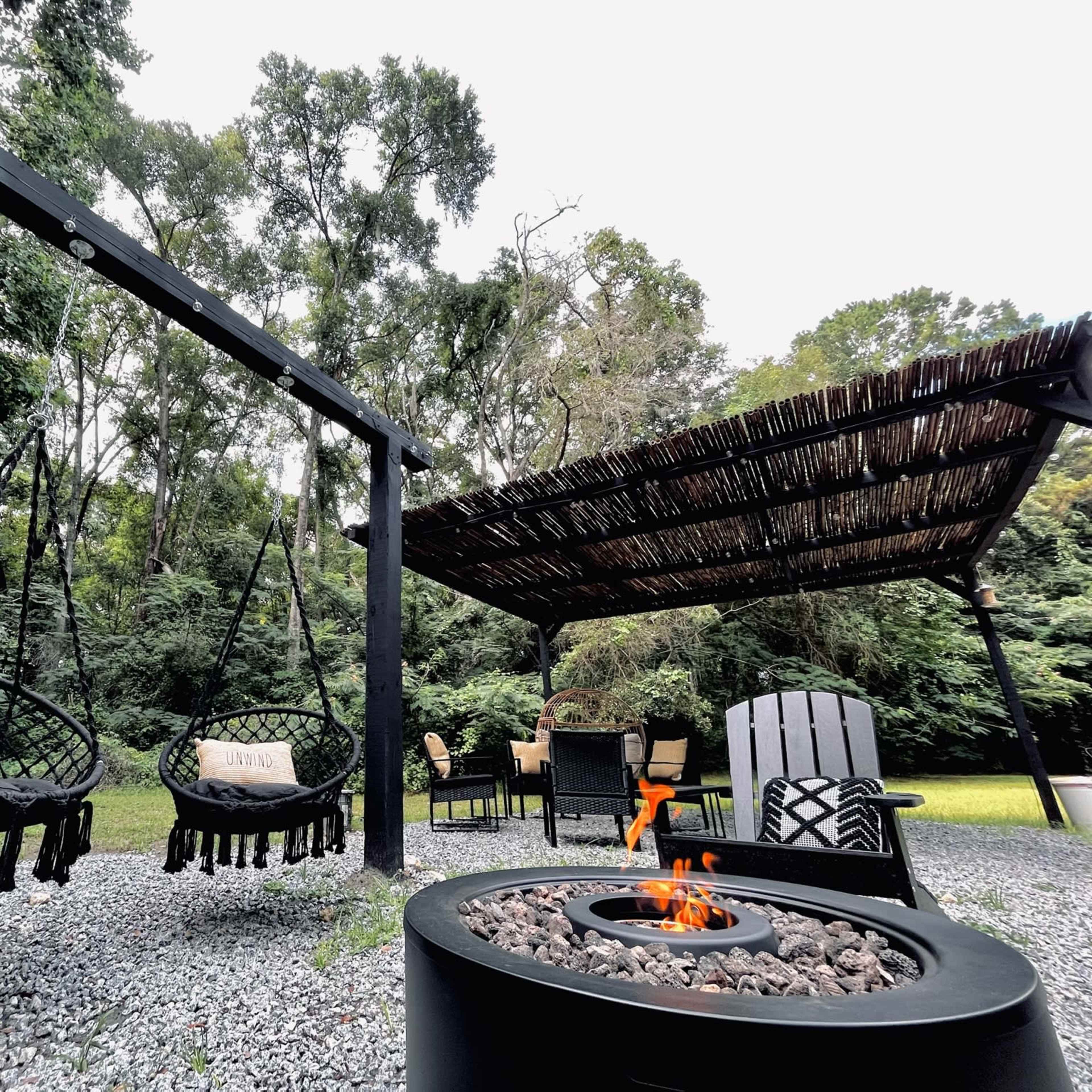 A outdoor patio area with two hanging chairs, a fire pit, and a covered seating space surrounded by trees.
