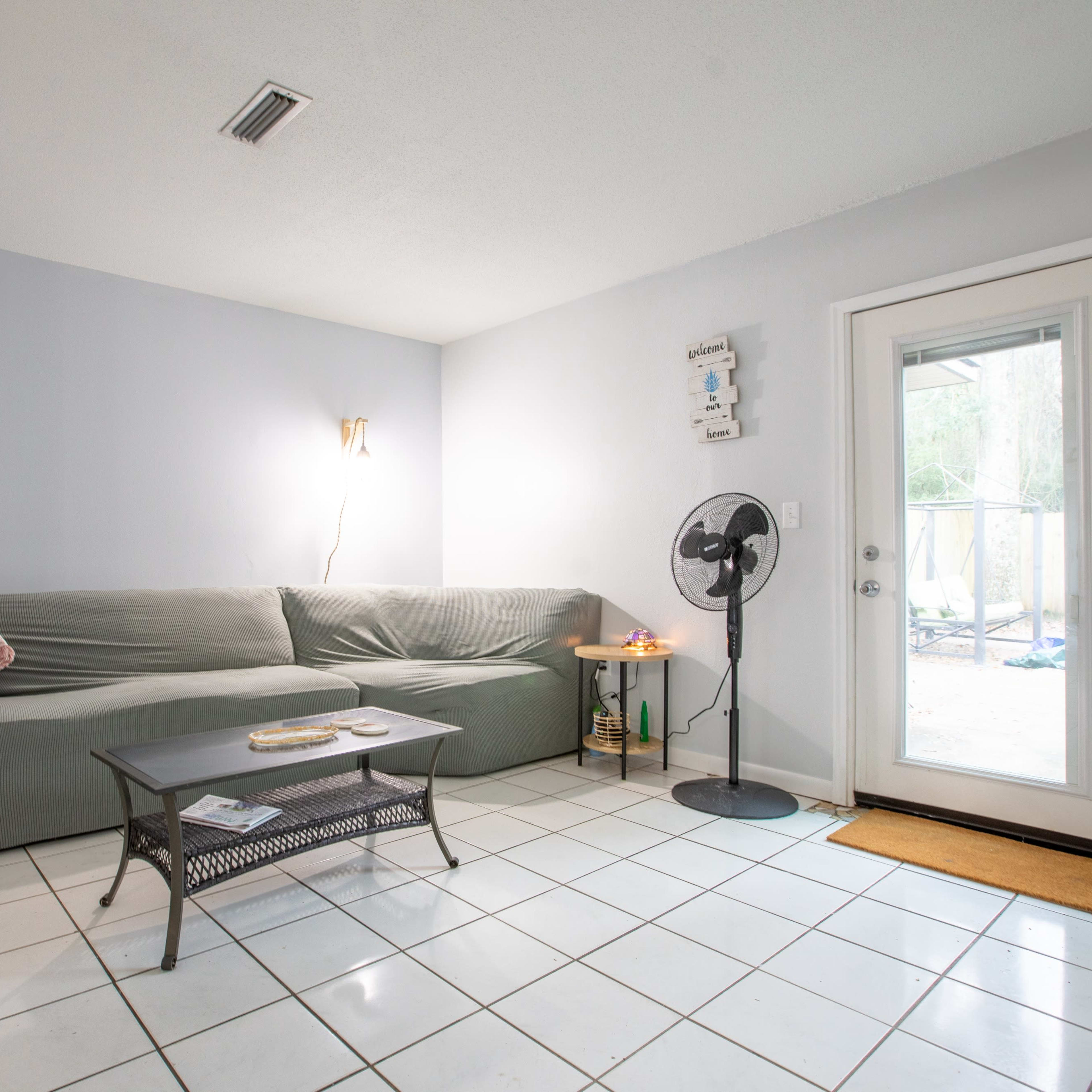 A light blue living room features a green sofa, a small table, a standing fan, and a door leading outside.