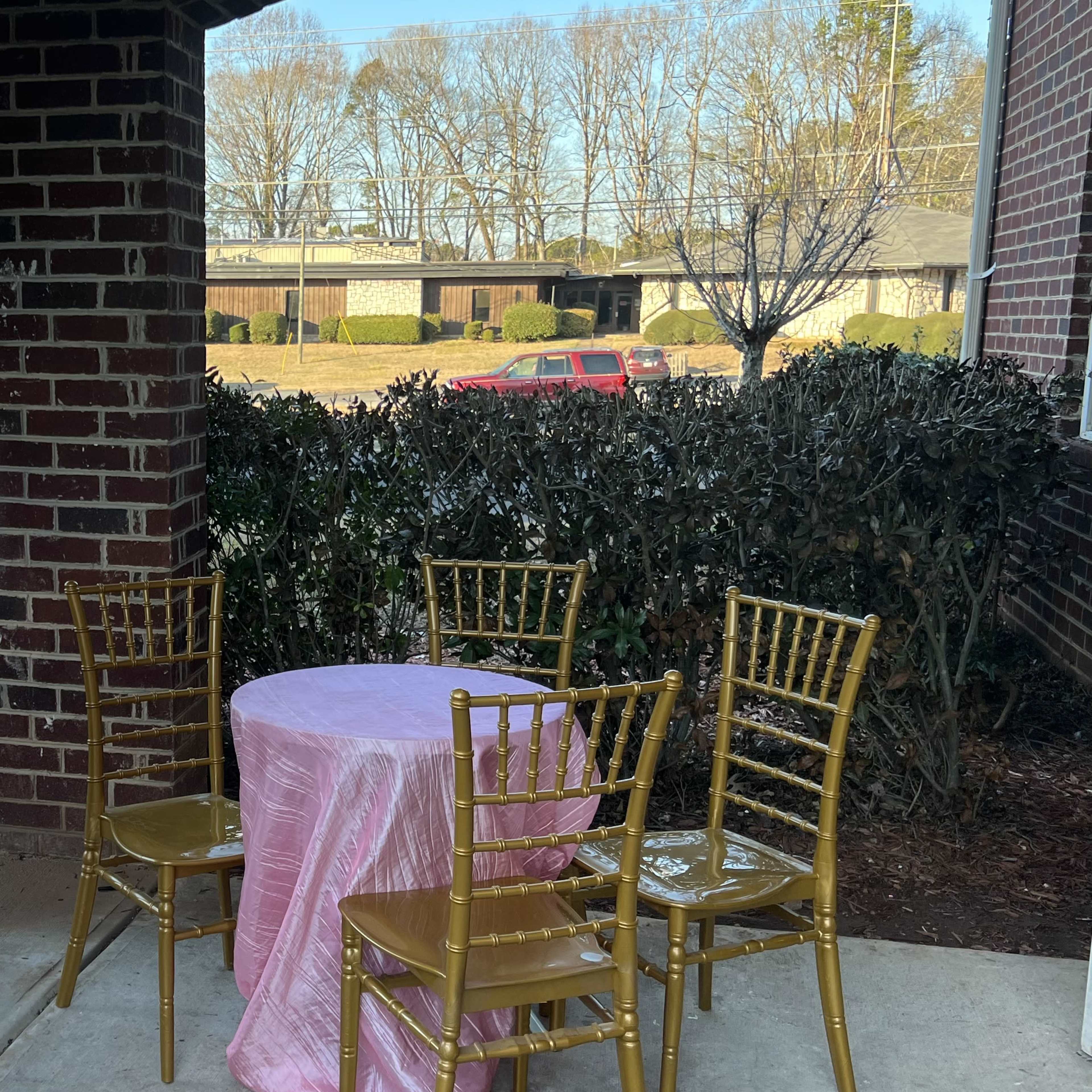 A round table covered with a pink tablecloth is set with four gold chairs in an outdoor area framed by brick columns and greenery.