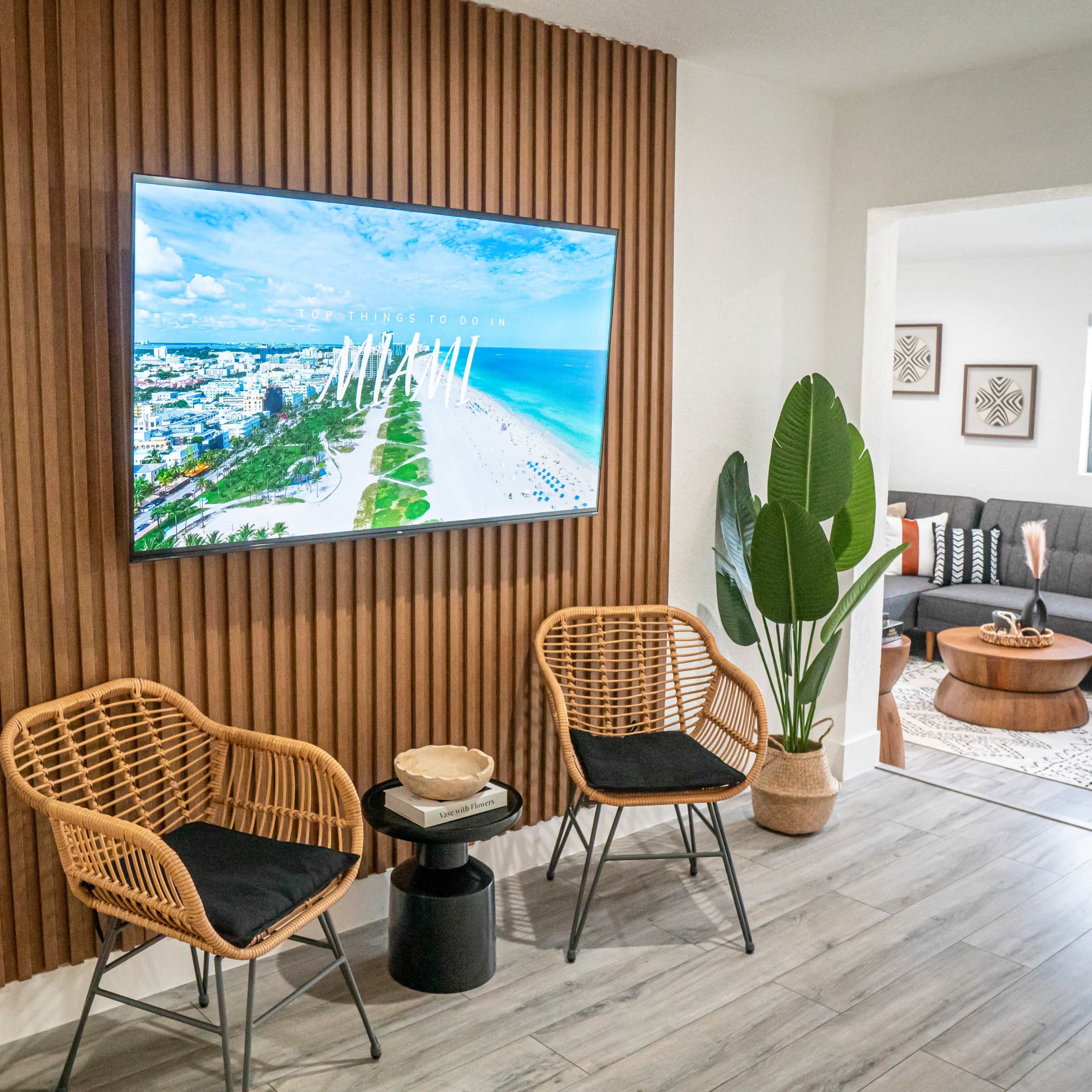 A modern living space with a wall-mounted television, two rattan chairs, and potted plants beside a cozy seating area featuring a coffee table and decorative accents.