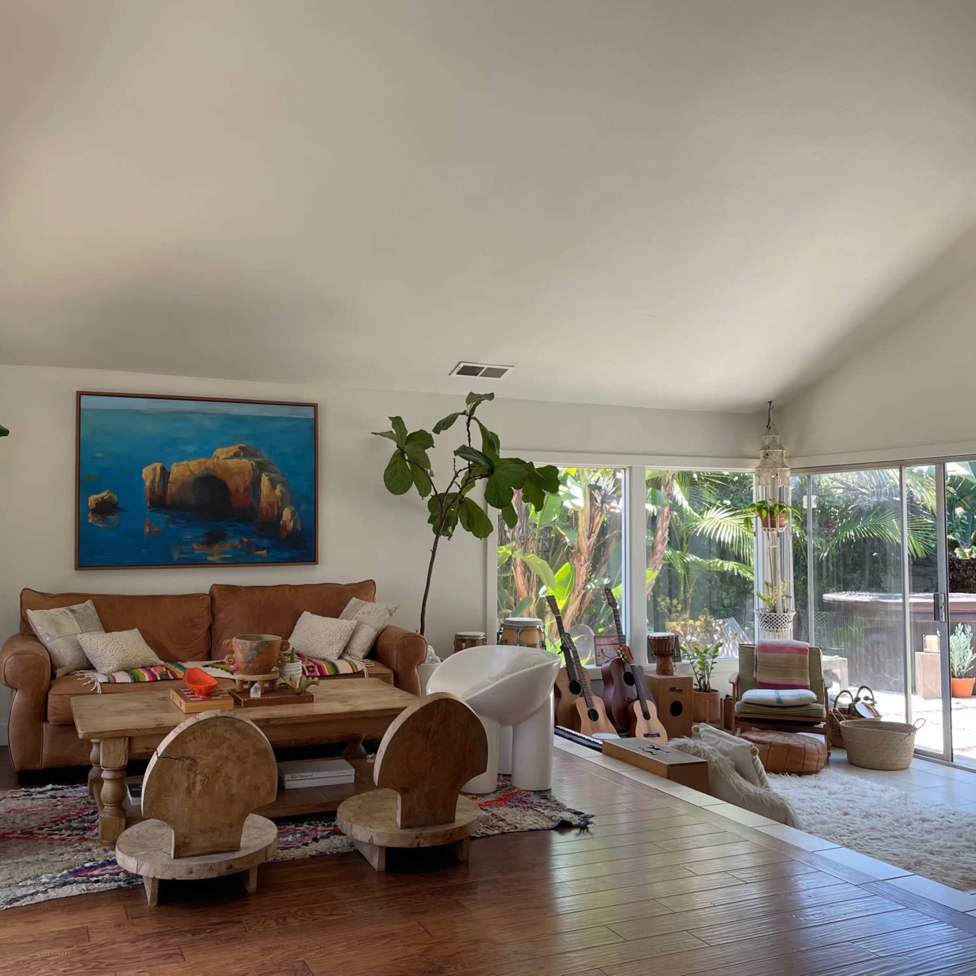 A living room with a large brown sofa, a wooden table, and various plants, surrounded by large windows that offer a view of the outdoors.