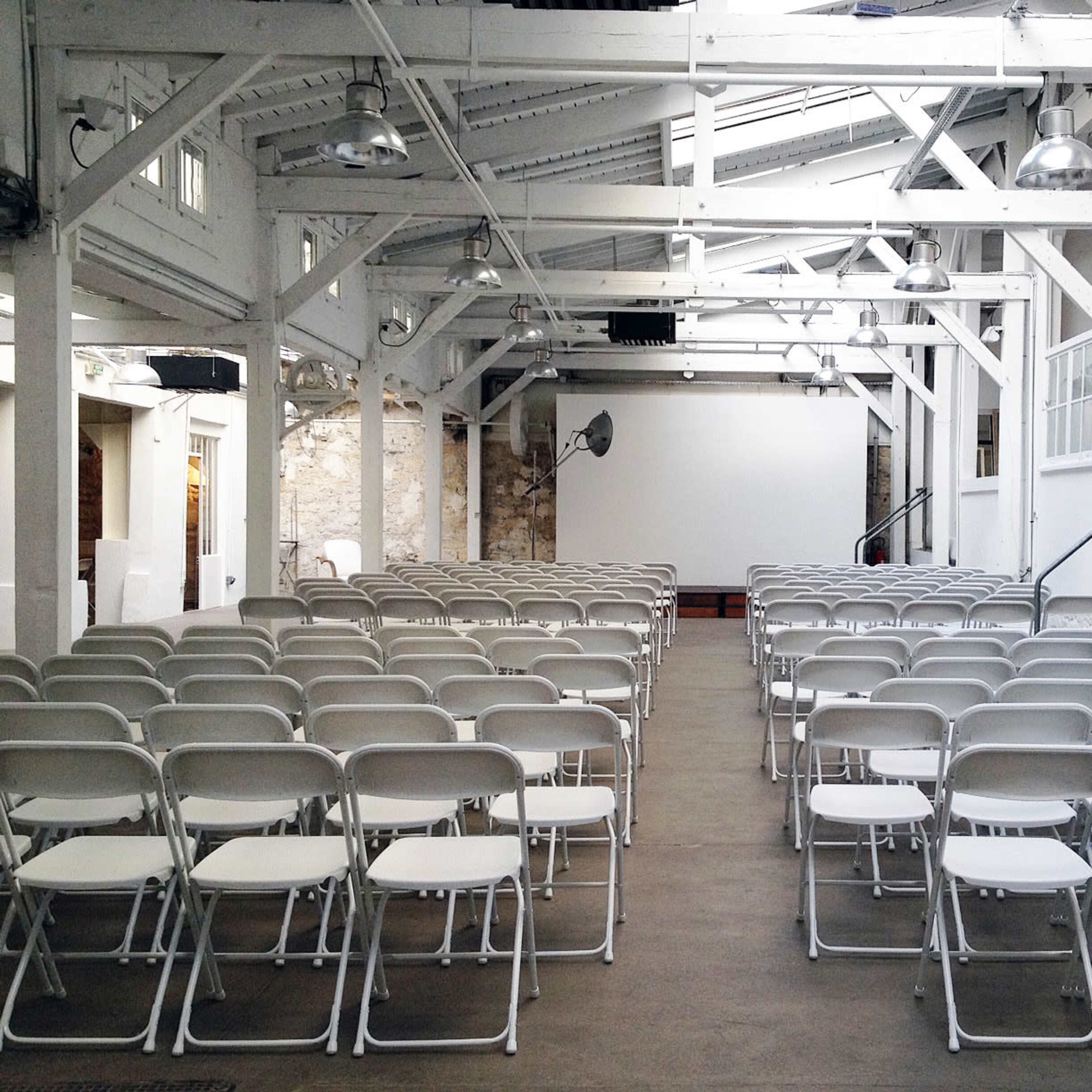 A venue features rows of empty white chairs facing a large projection screen in a bright, industrial space.