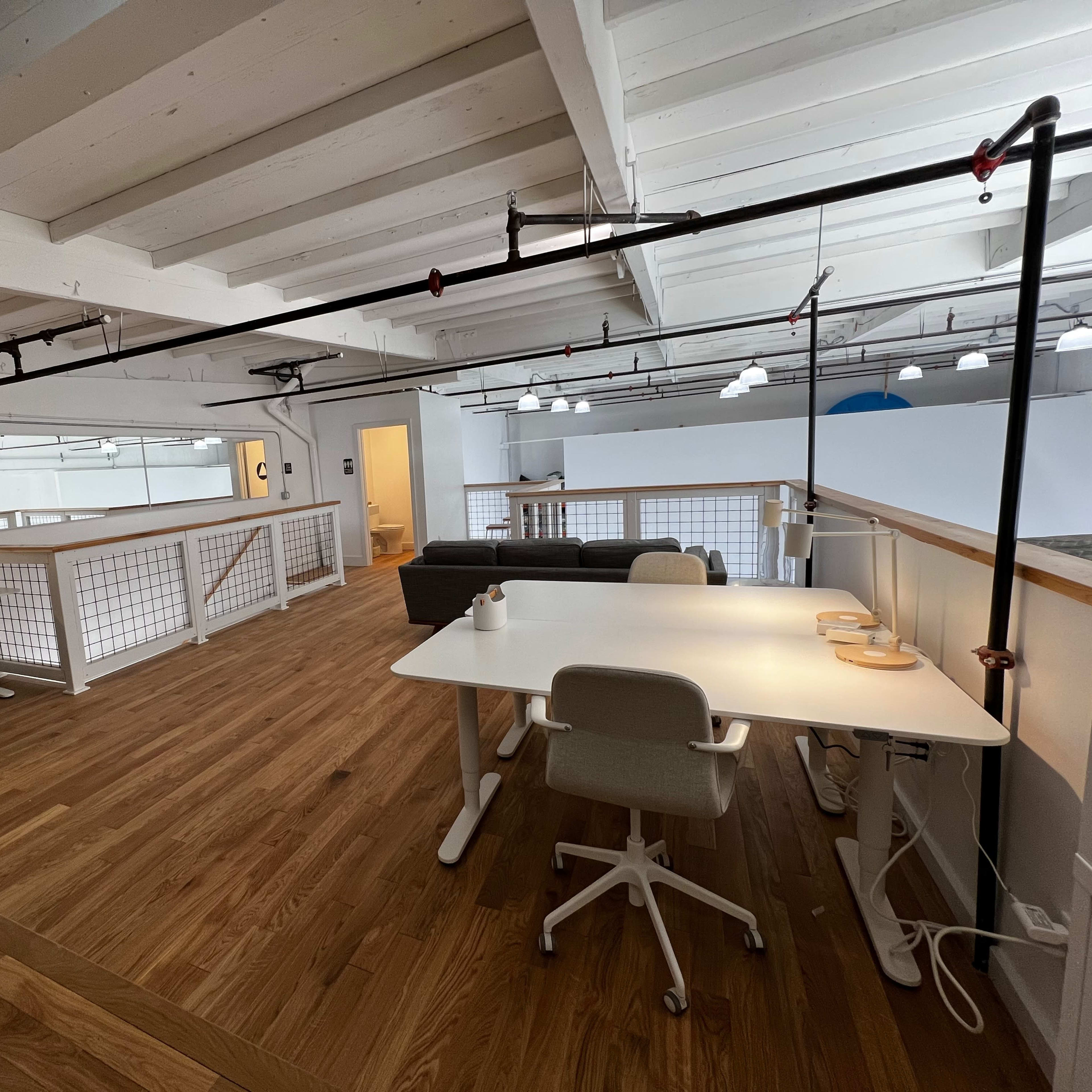 A spacious, modern office interior with wooden flooring, two workstations, a sofa, and overhead lighting.