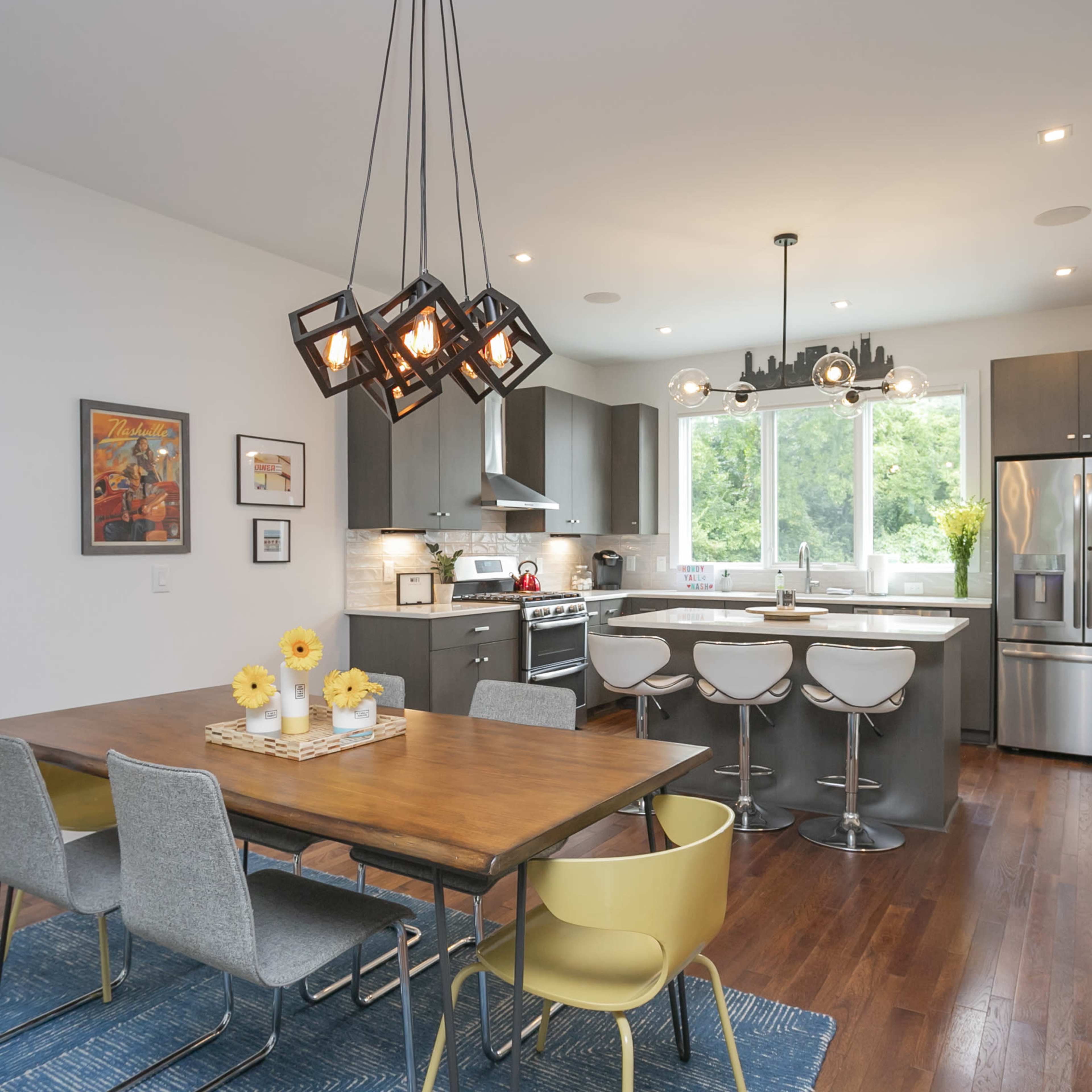 A modern kitchen and dining area features a wooden table surrounded by chairs, with a sleek kitchen counter, stainless steel appliances, and large windows providing natural light.