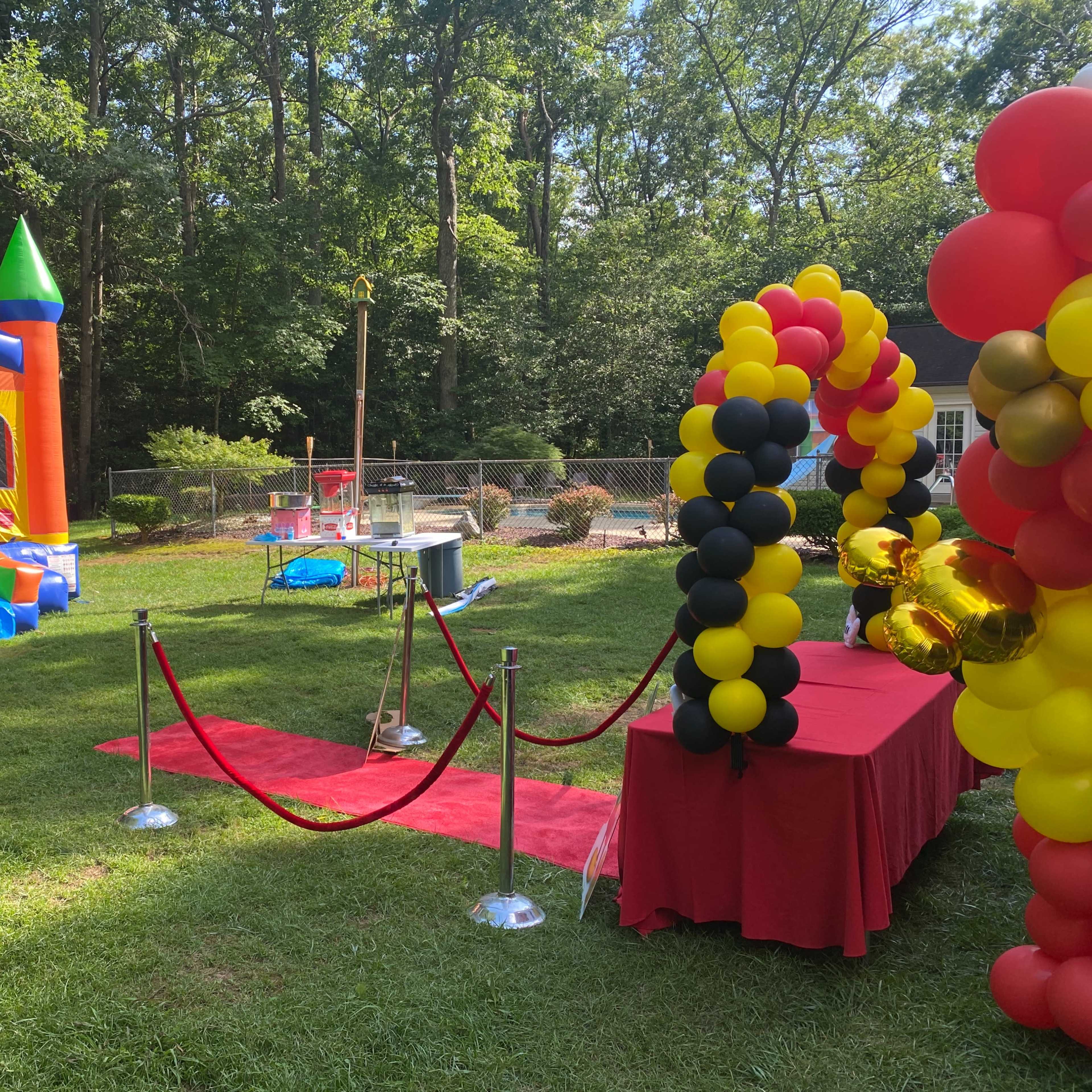 The scene features a colorful bounce house, a red carpet with stanchions, a table draped in red fabric, and a background of greenery and playground equipment in a backyard.
