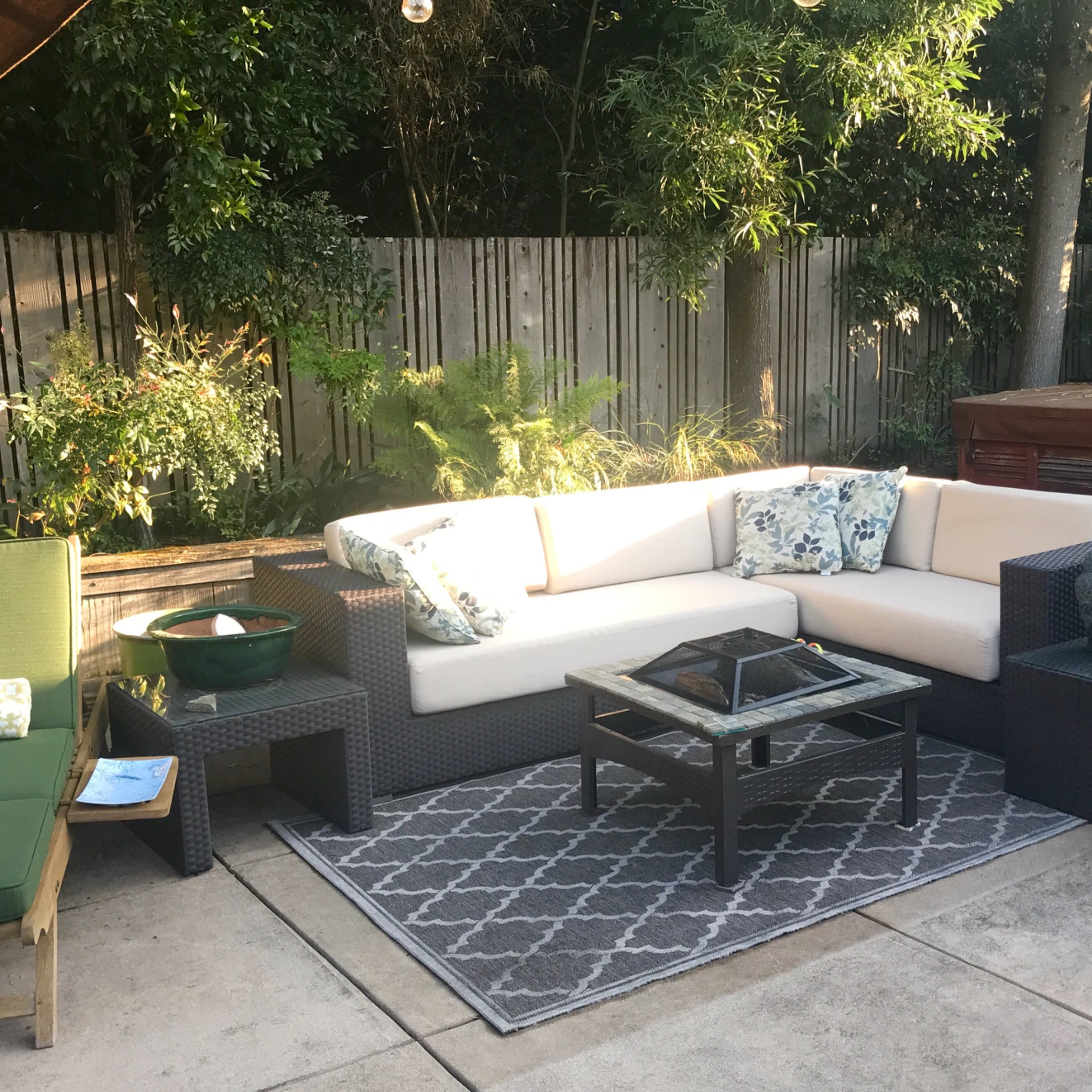 A landscaped outdoor seating area featuring a sectional sofa with cushions, a glass coffee table, and various plants in pots and raised beds.