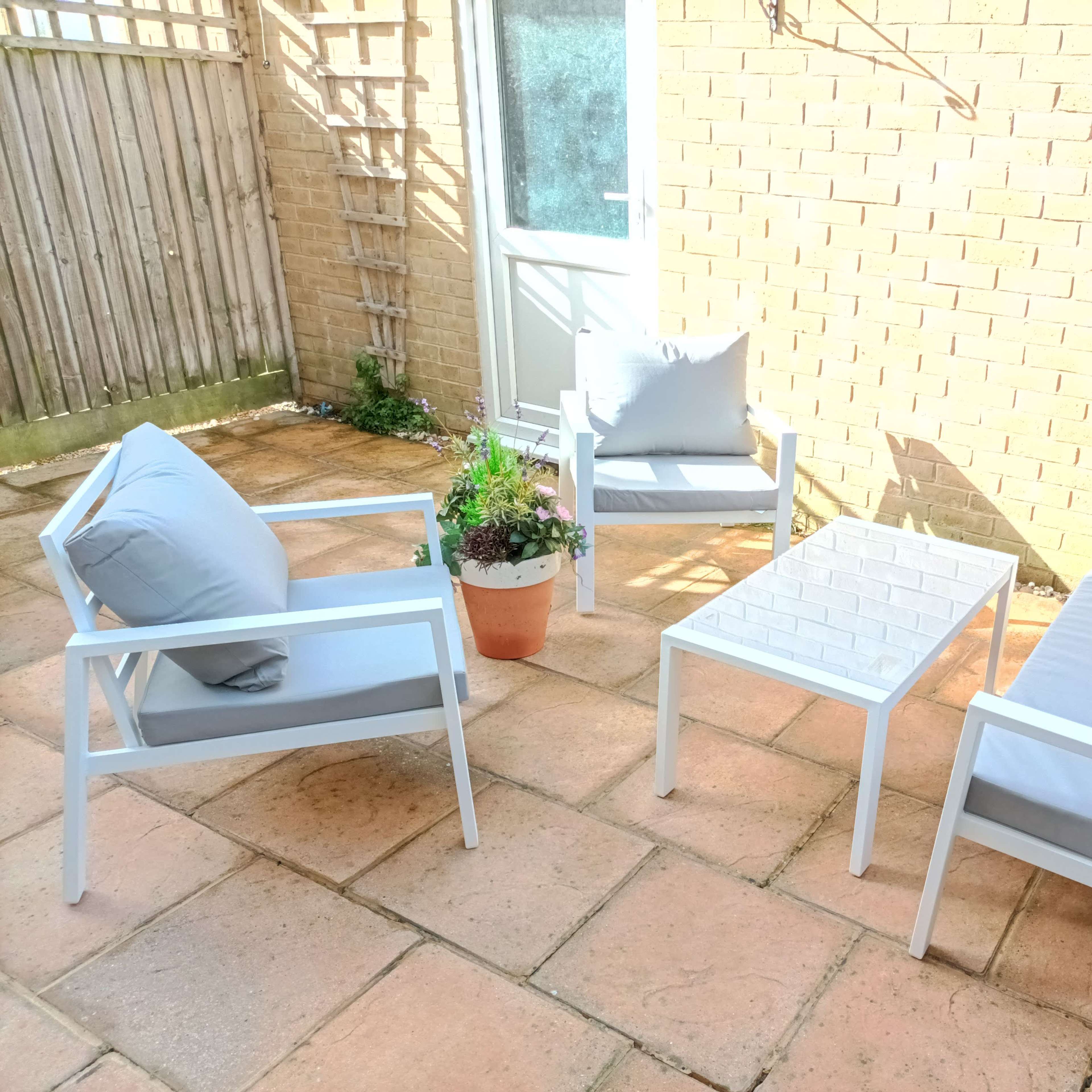 The scene shows a small outdoor patio area with four white metal-framed chairs, a matching rectangular table, and a potted plant, all set on stone tiles against a brick wall.