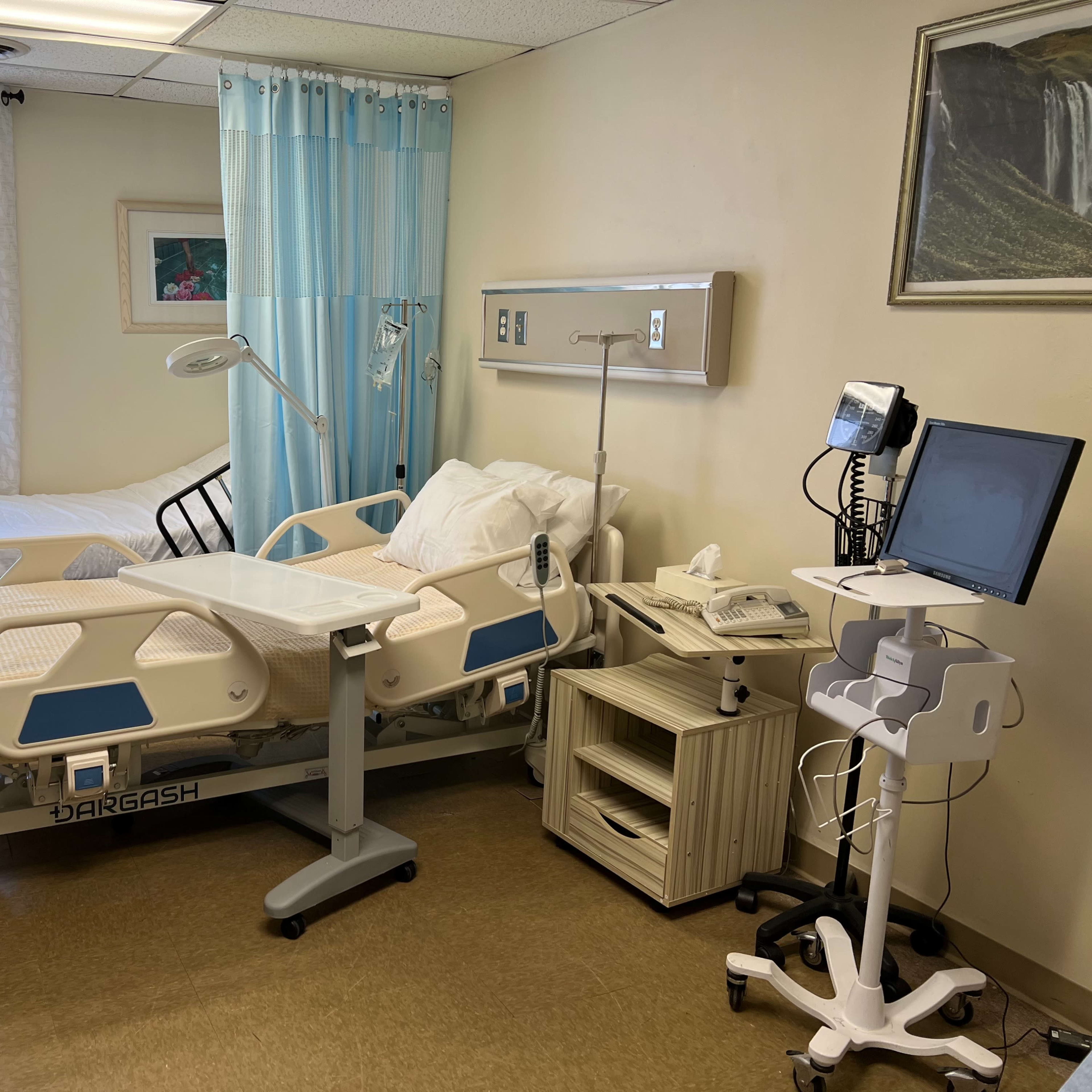 A hospital room with two beds, a bedside table, a medical monitor, and a computer on a mobile stand.