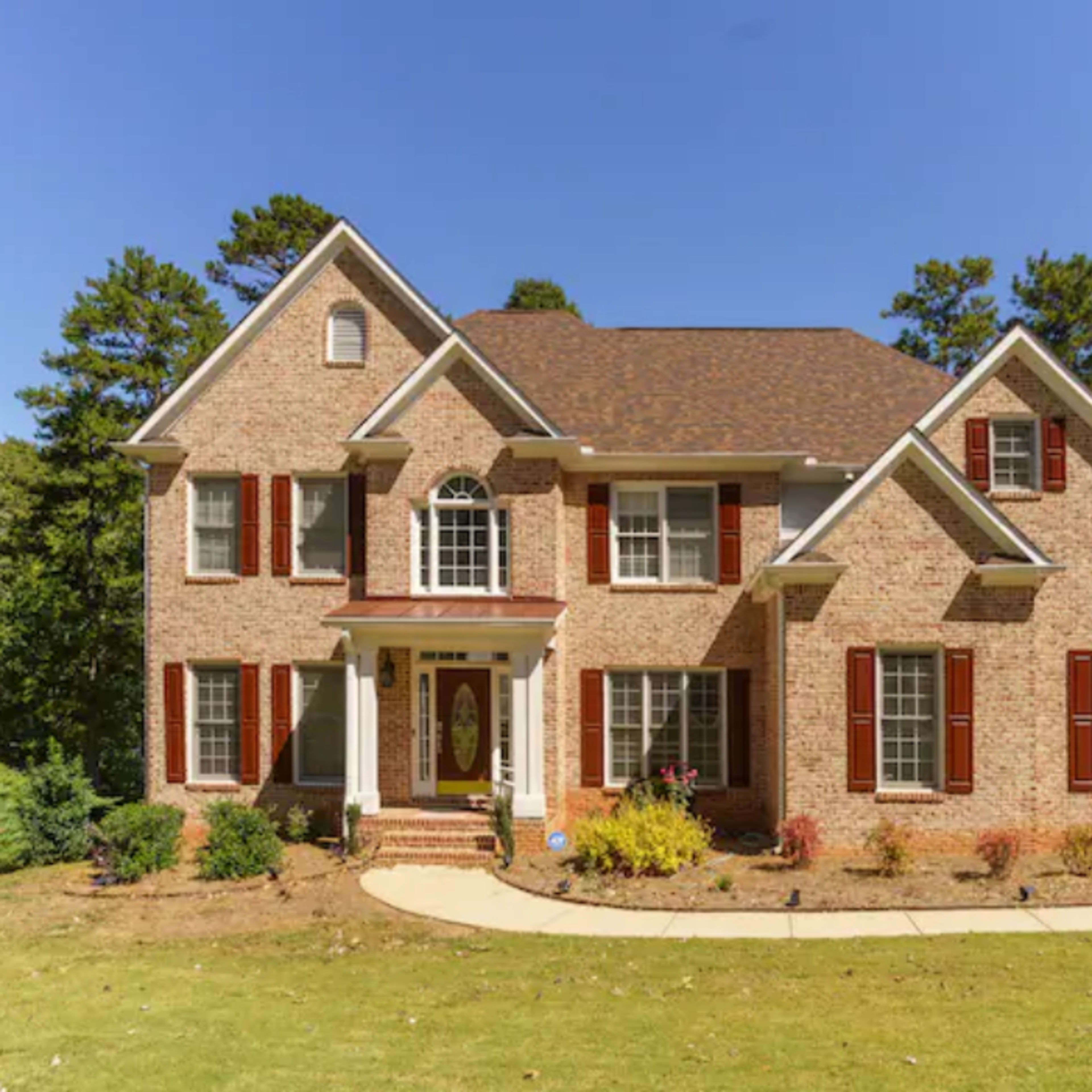 A two-story brick house with a symmetrical facade, featuring white trim and red shutters, surrounded by landscaped greenery.