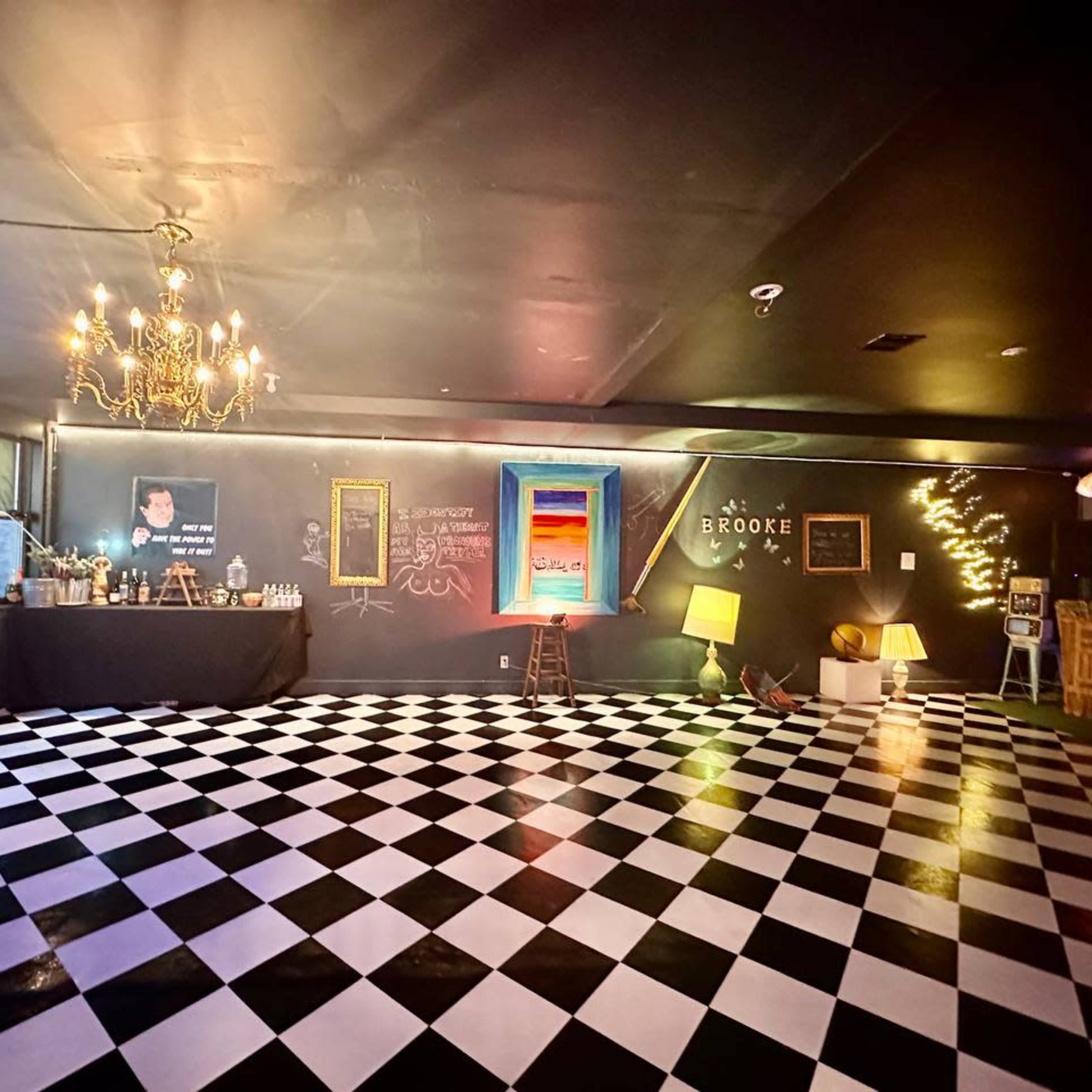 An empty room with a black and white checkered floor, a chandelier, a bar area, and a chalkboard wall with colorful artwork and text.