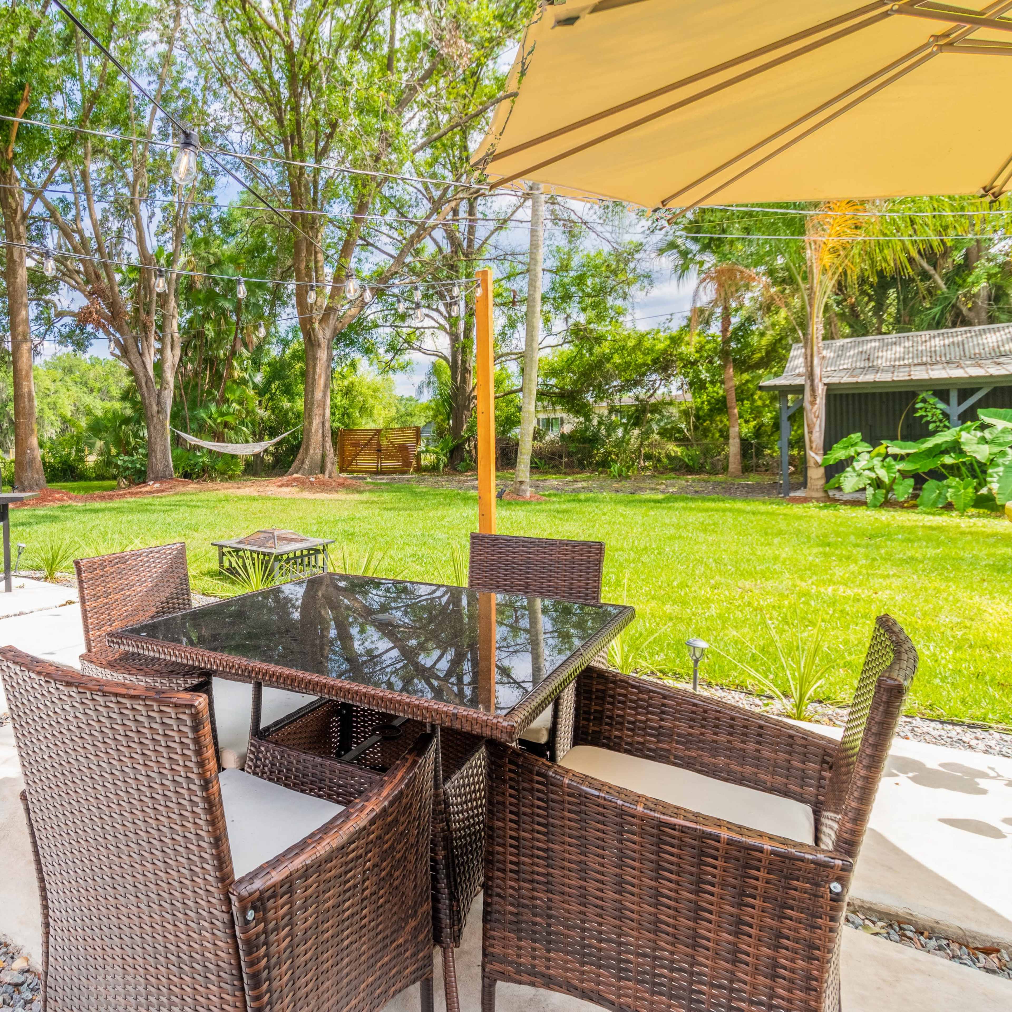 A patio area features a black table surrounded by wicker chairs, with a grill nearby and a lush green yard in the background.