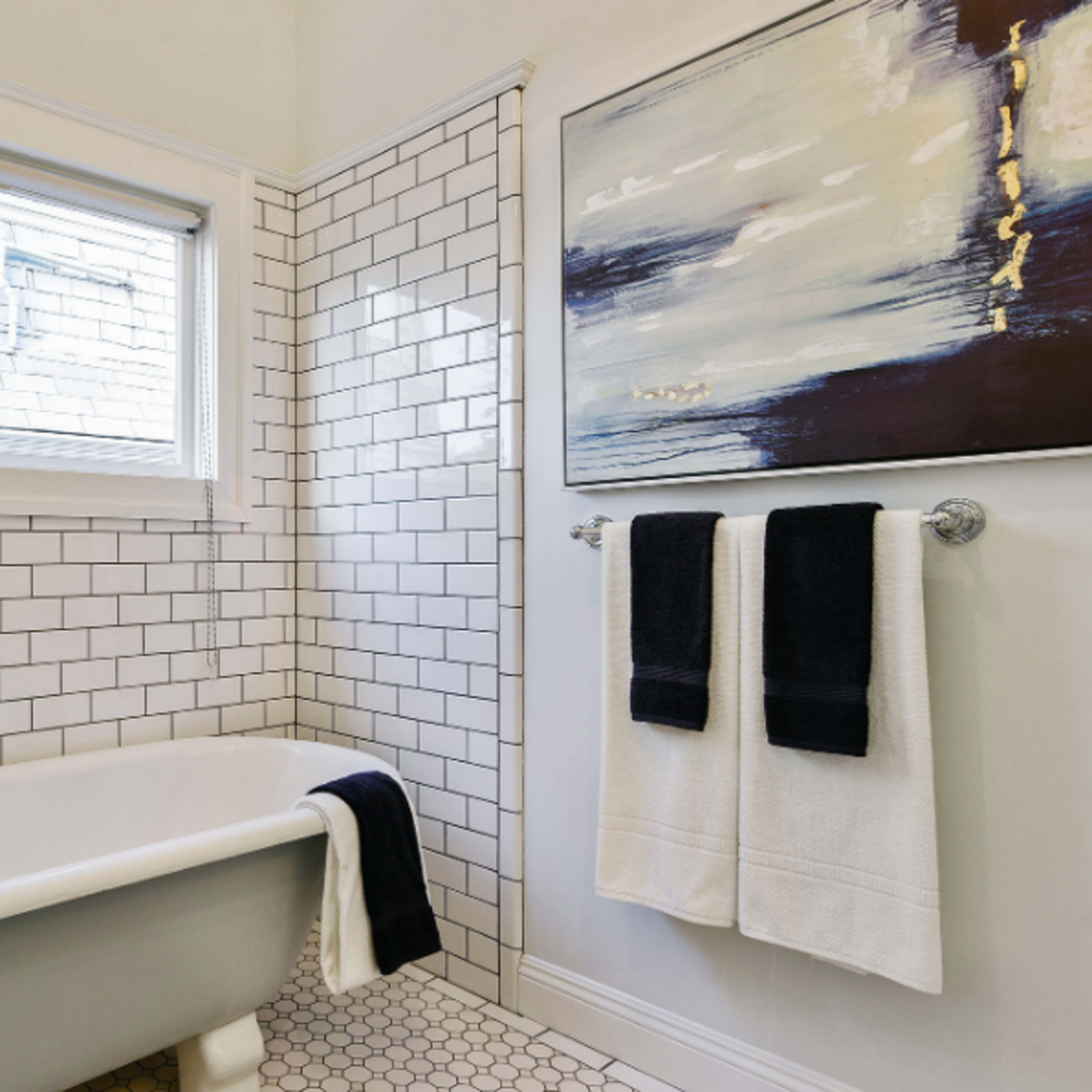 A bathroom featuring a freestanding white bathtub, white subway tile walls, and a framed abstract painting, with two towels hanging on a rack.