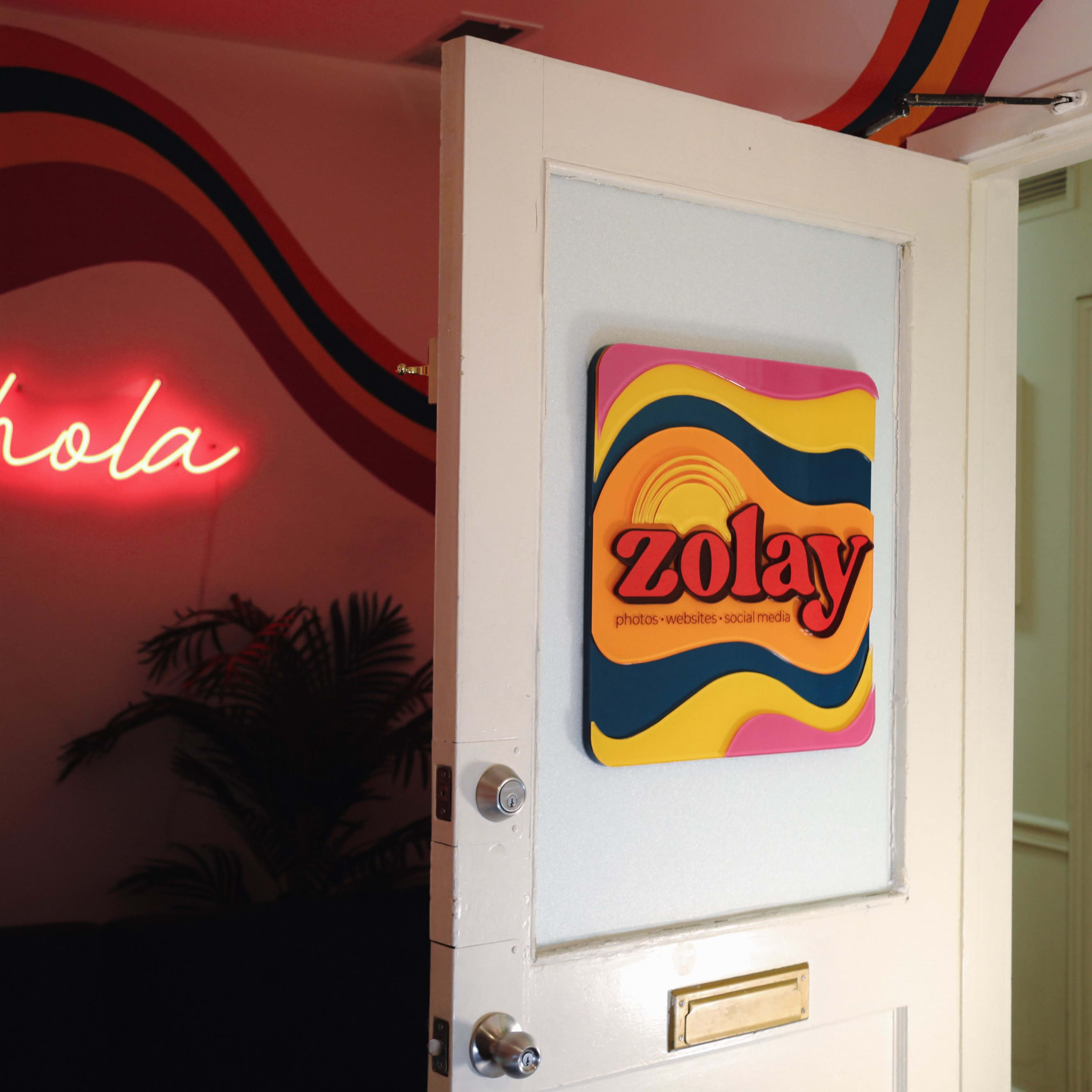A door partially open reveals a colorful wall with the words "hola" in neon and a sign that reads "zolay" on the adjacent wall.