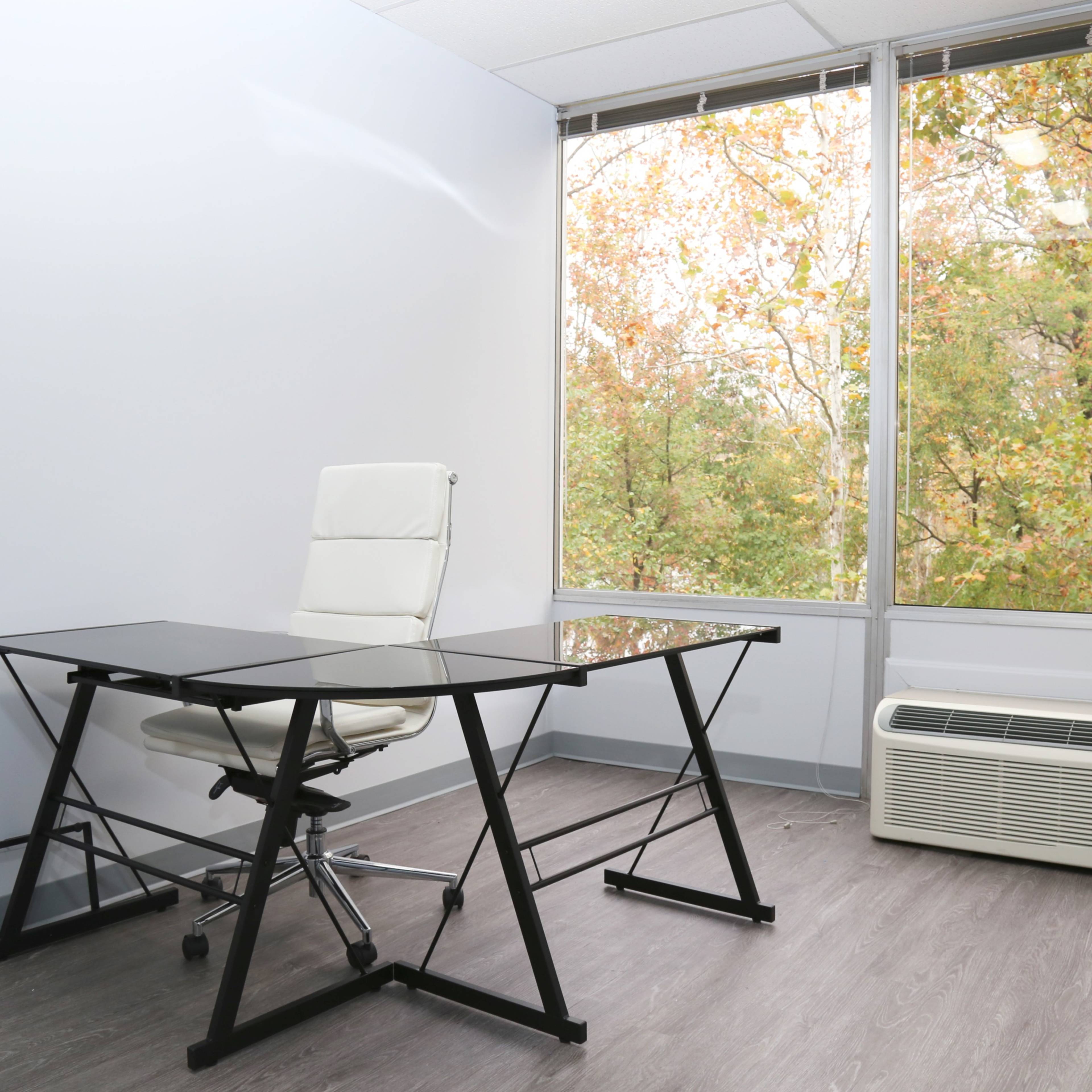 A modern office features a glass-topped desk, a white ergonomic chair, and large windows displaying autumn foliage.