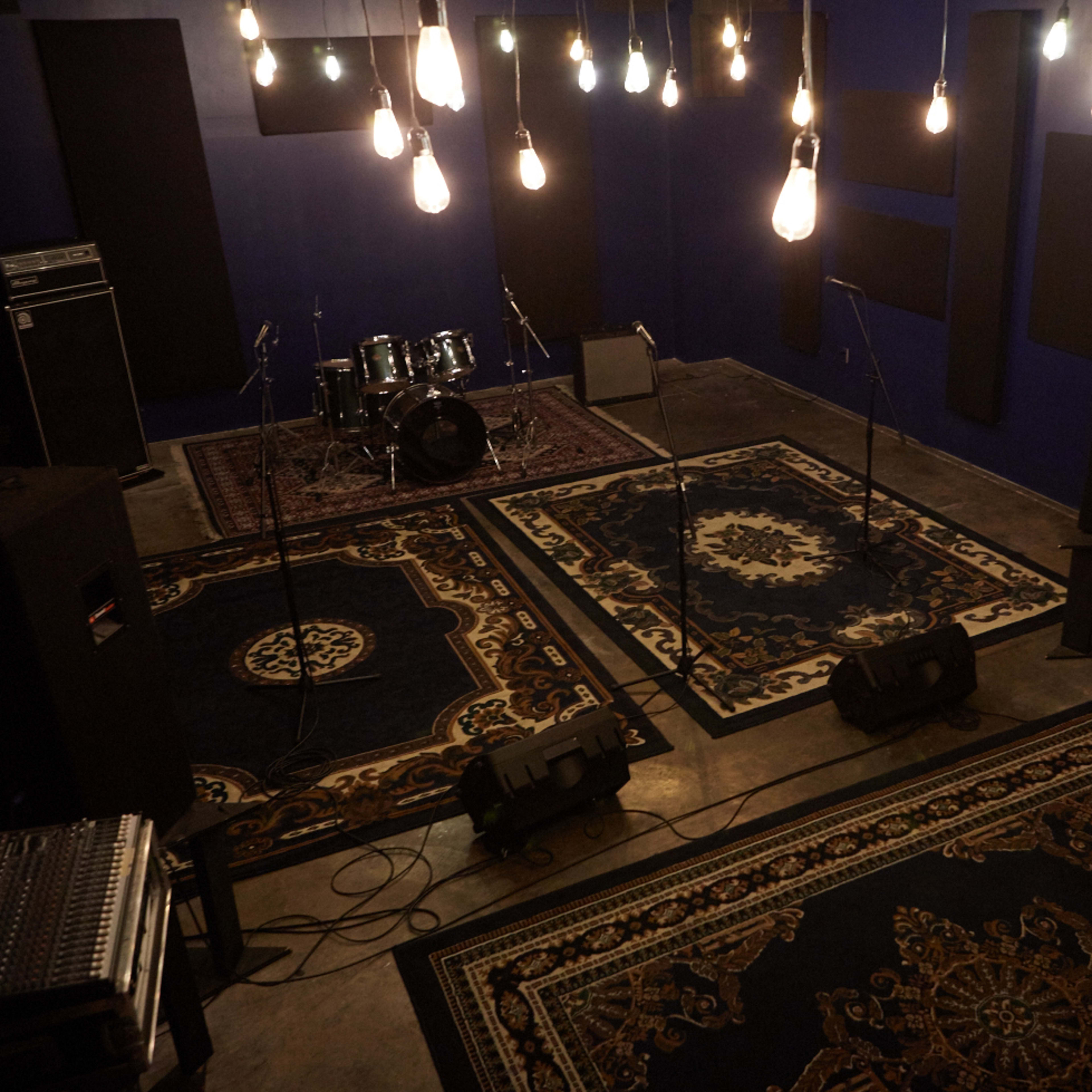 A music rehearsal space features a drum set, microphones on stands, and illuminated by hanging light bulbs, with rugs on a concrete floor.