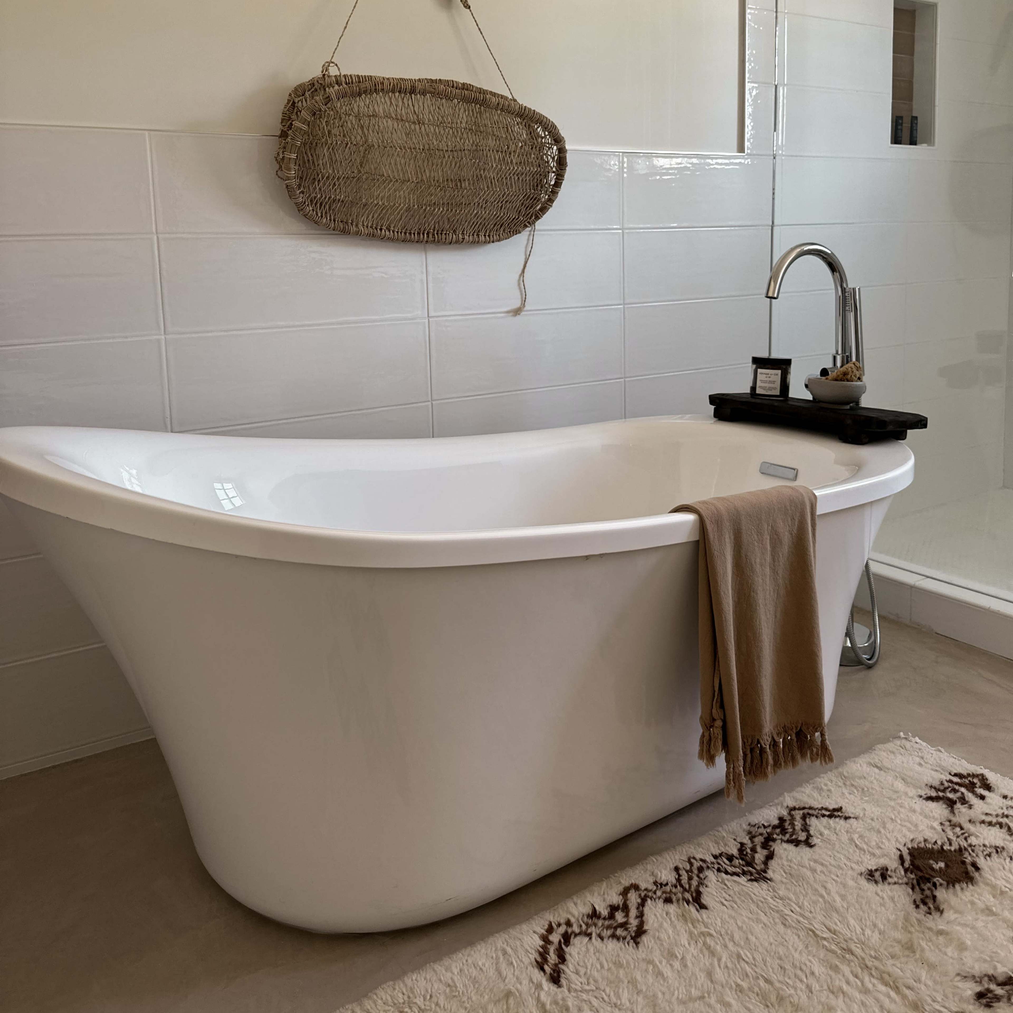 A modern bathroom featuring a freestanding white bathtub, a woven wall hanging, and a wooden shelf with a candle beside the tub.