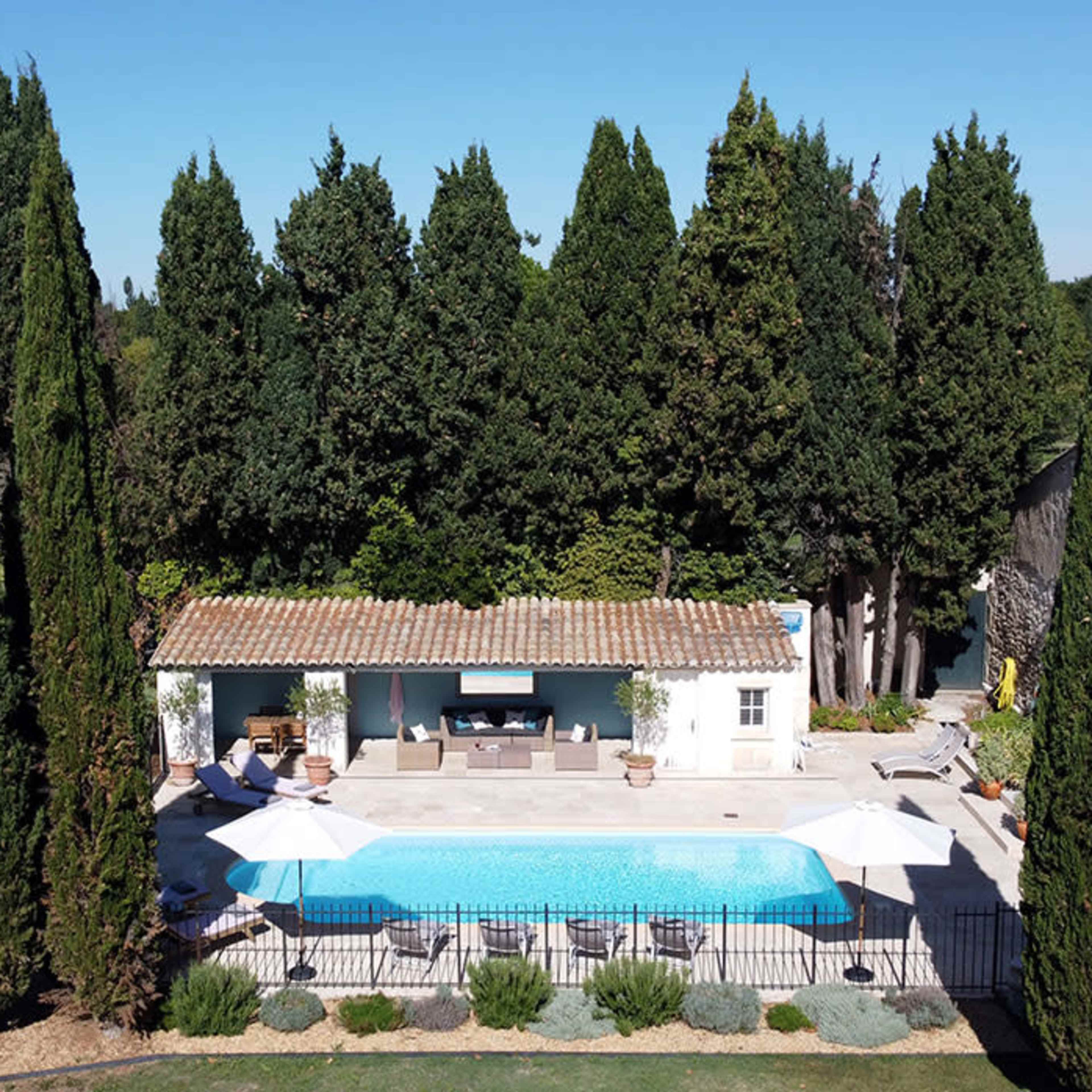 A rectangular swimming pool is surrounded by tall cypress trees and a stone patio with lounge chairs and umbrellas.