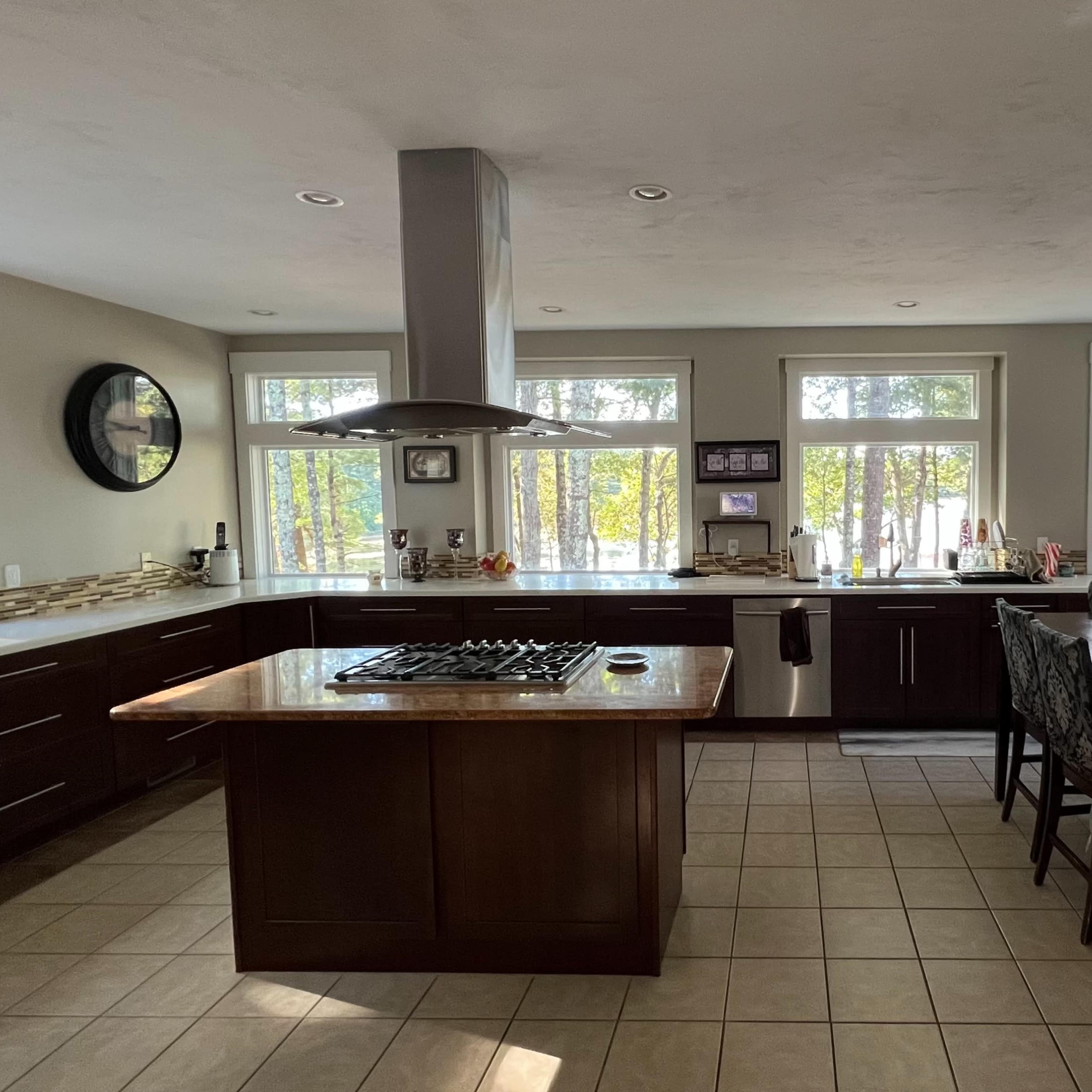 A modern kitchen features dark wood cabinets, an island with a gas stove, and large windows overlooking a wooded area.